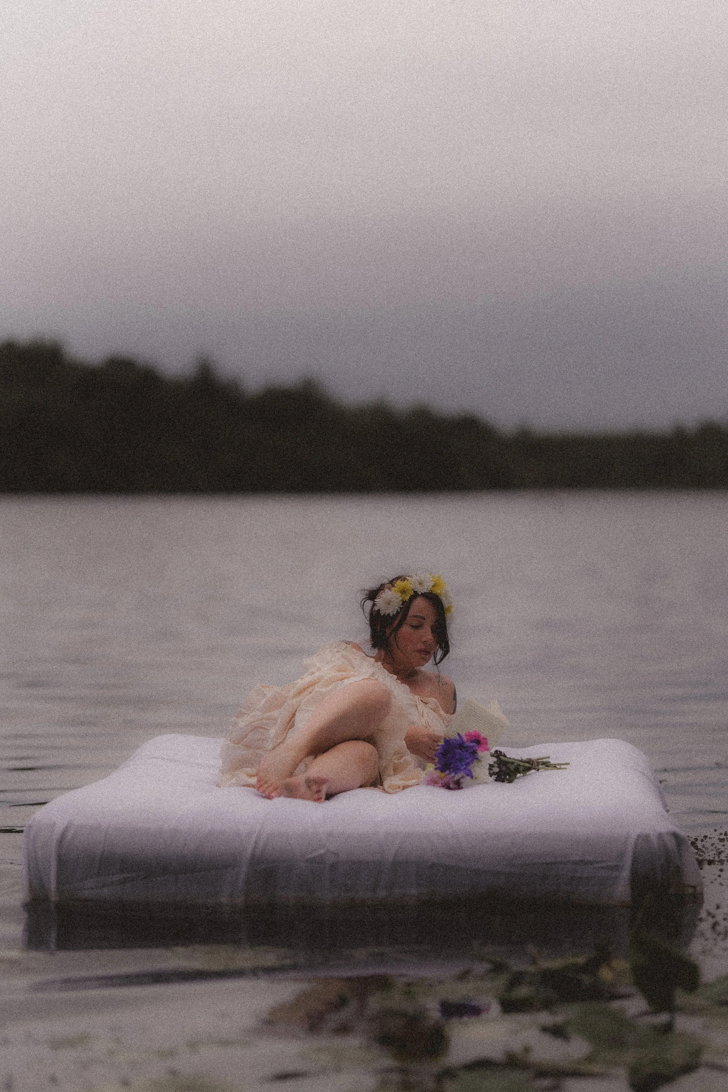 A woman sitting on a blanket on the water with a floral crown on her head, holding flowers and looking down, near a body of water with trees in the background.