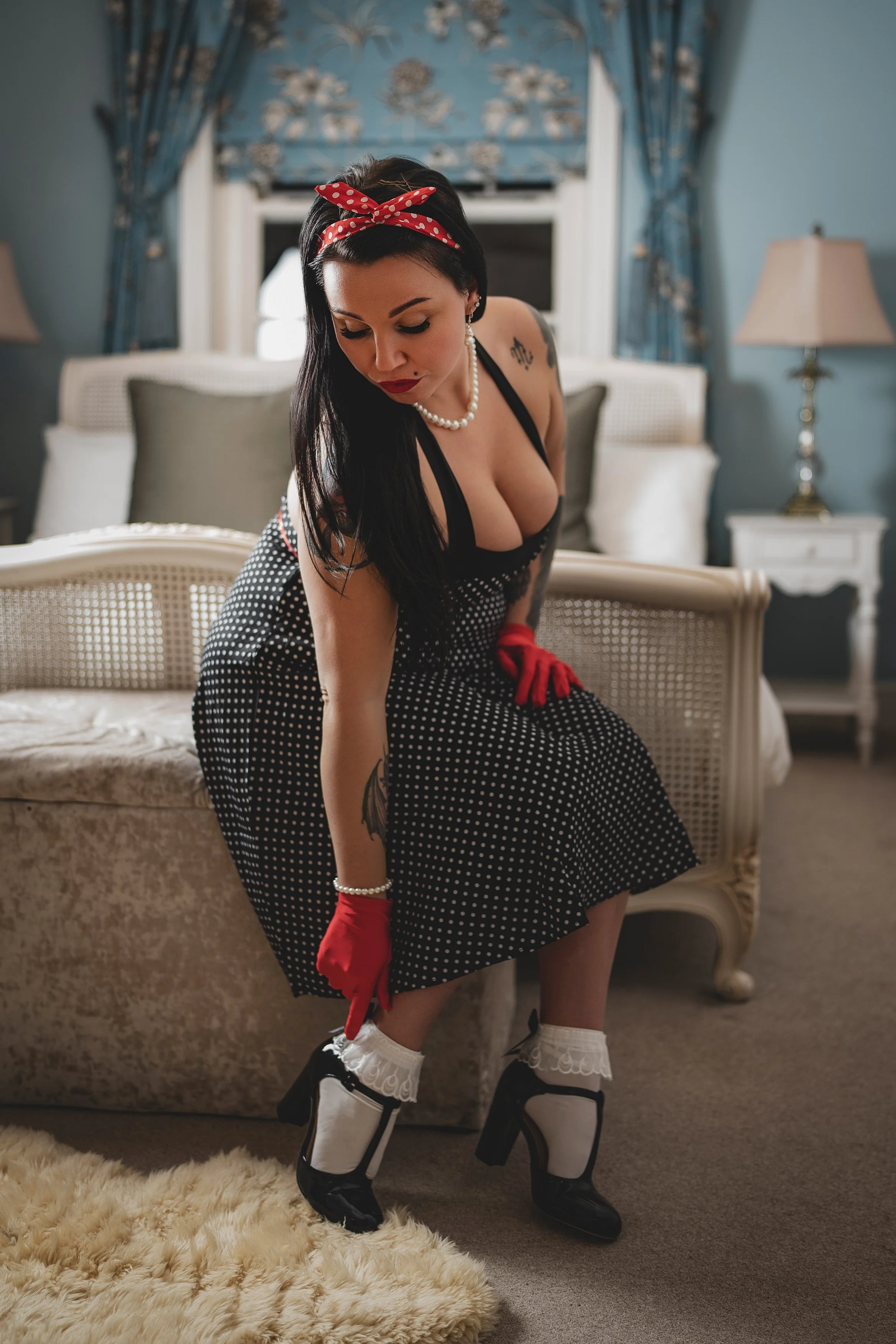 A woman in vintage pin-up style clothing, wearing a polka-dot dress, red gloves, and high heels, is adjusting her sock in a well-decorated bedroom.