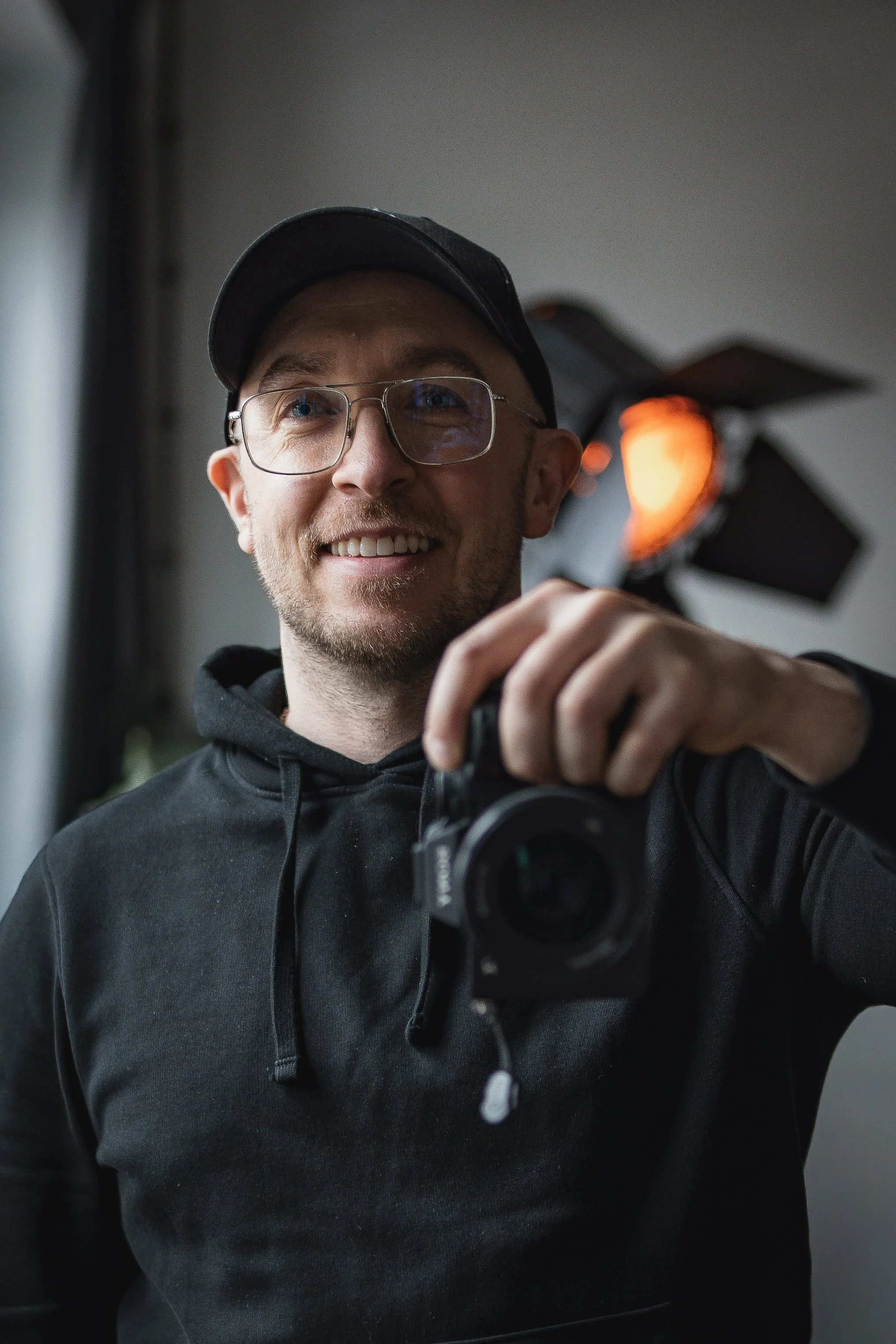 A man with glasses and a black hoodie smiling and taking a selfie with a camera, with a large studio light in the background.
