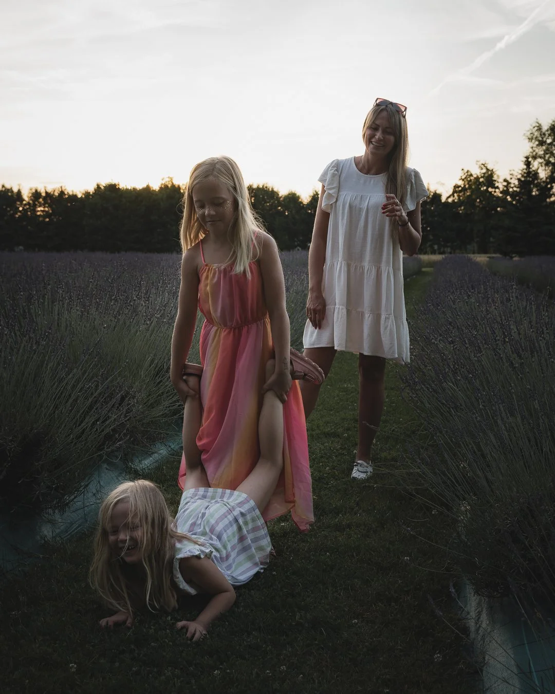 A woman and three young girls in a lavender field during sunset, with two girls standing and one girl lying on the ground, partially wrapped in a blanket.