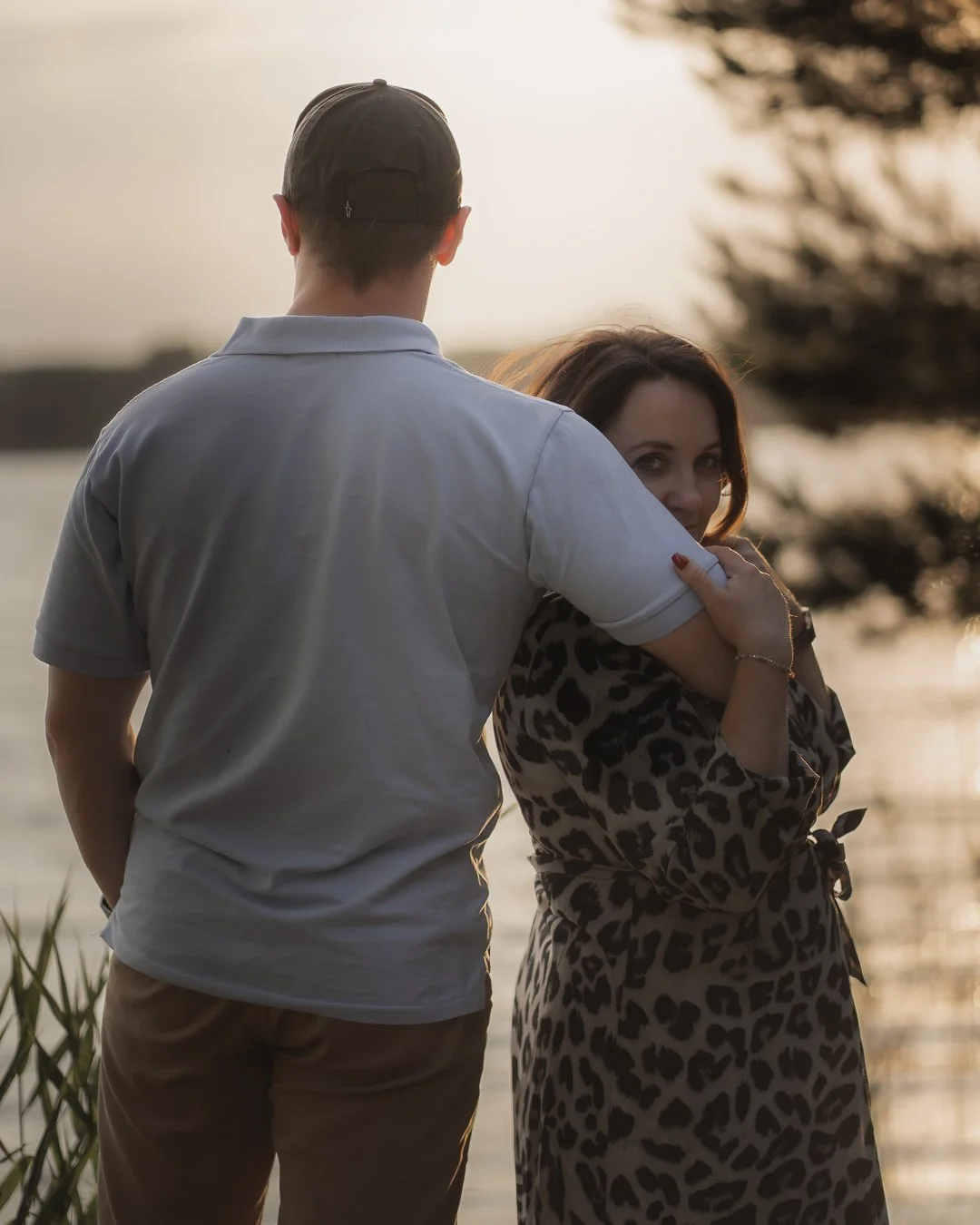 A man and a woman standing near a body of water at sunset, with the woman looking at the camera and the man facing away.