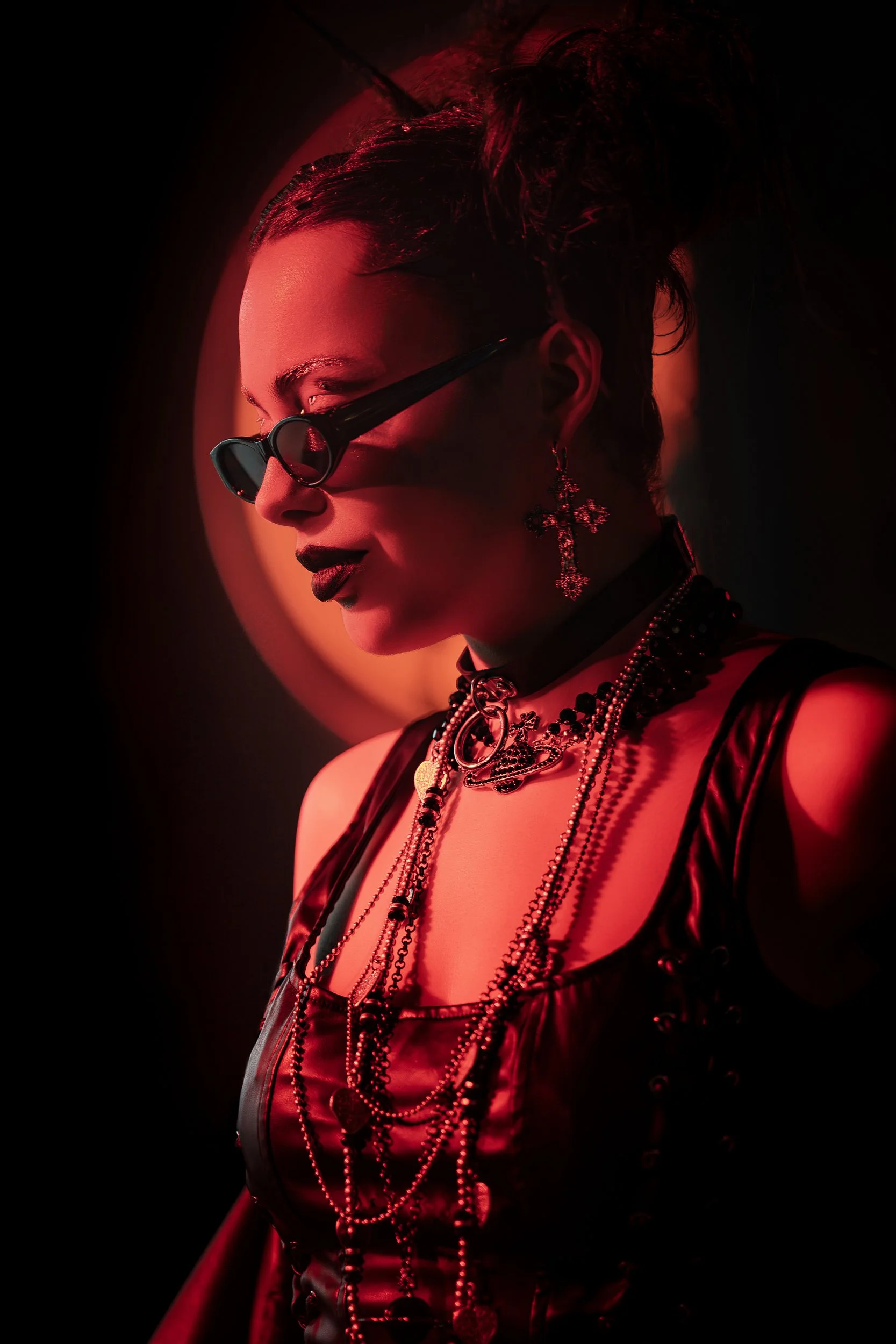 Side profile of a woman with dark lipstick, wearing dark sunglasses, and adorned with layered jewelry including necklaces, earrings, and a choker, illuminated by red lighting.