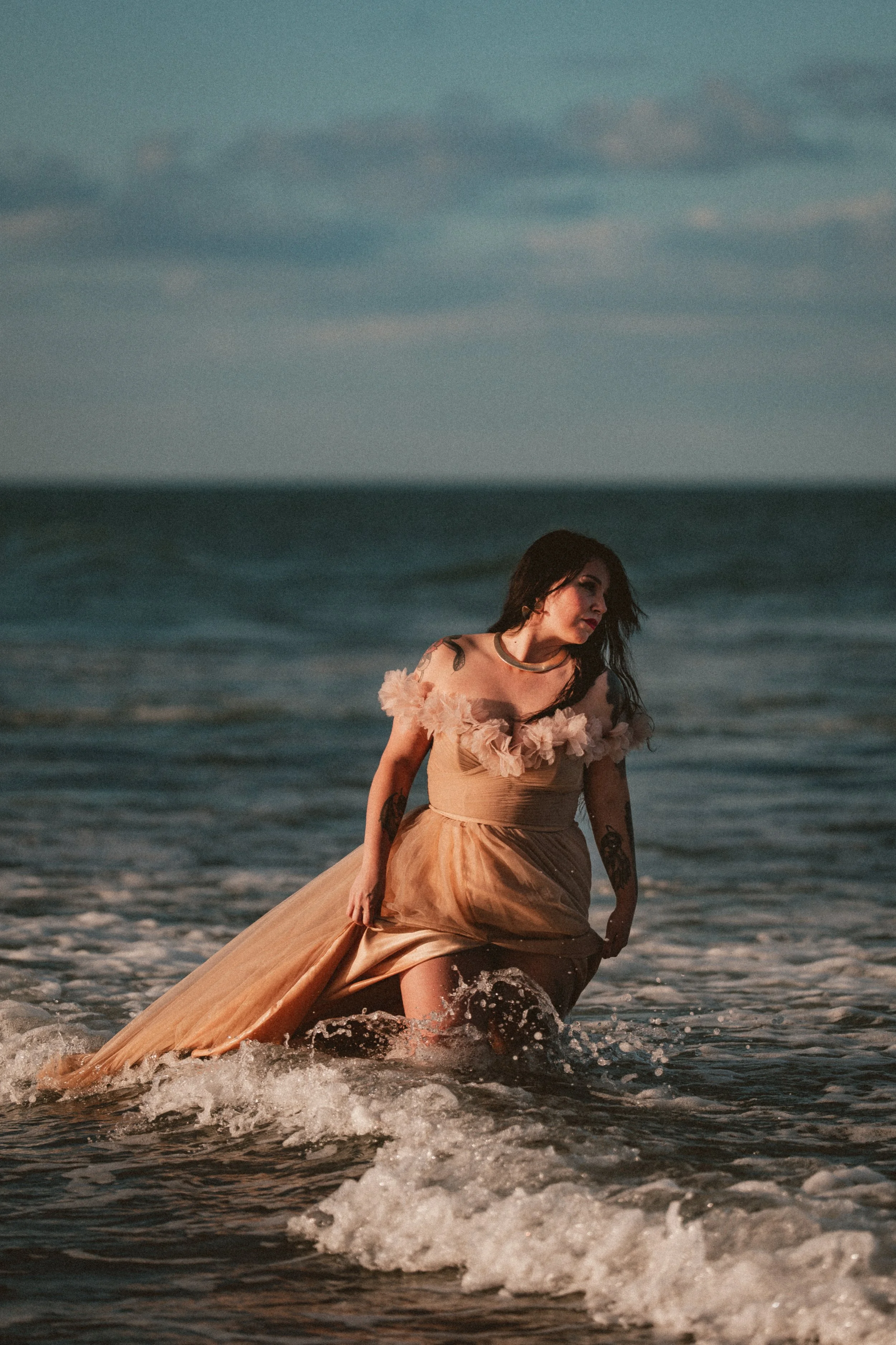 Woman in a flowing beige dress with floral details at the shoulder, kneeling in the ocean waves at sunset.