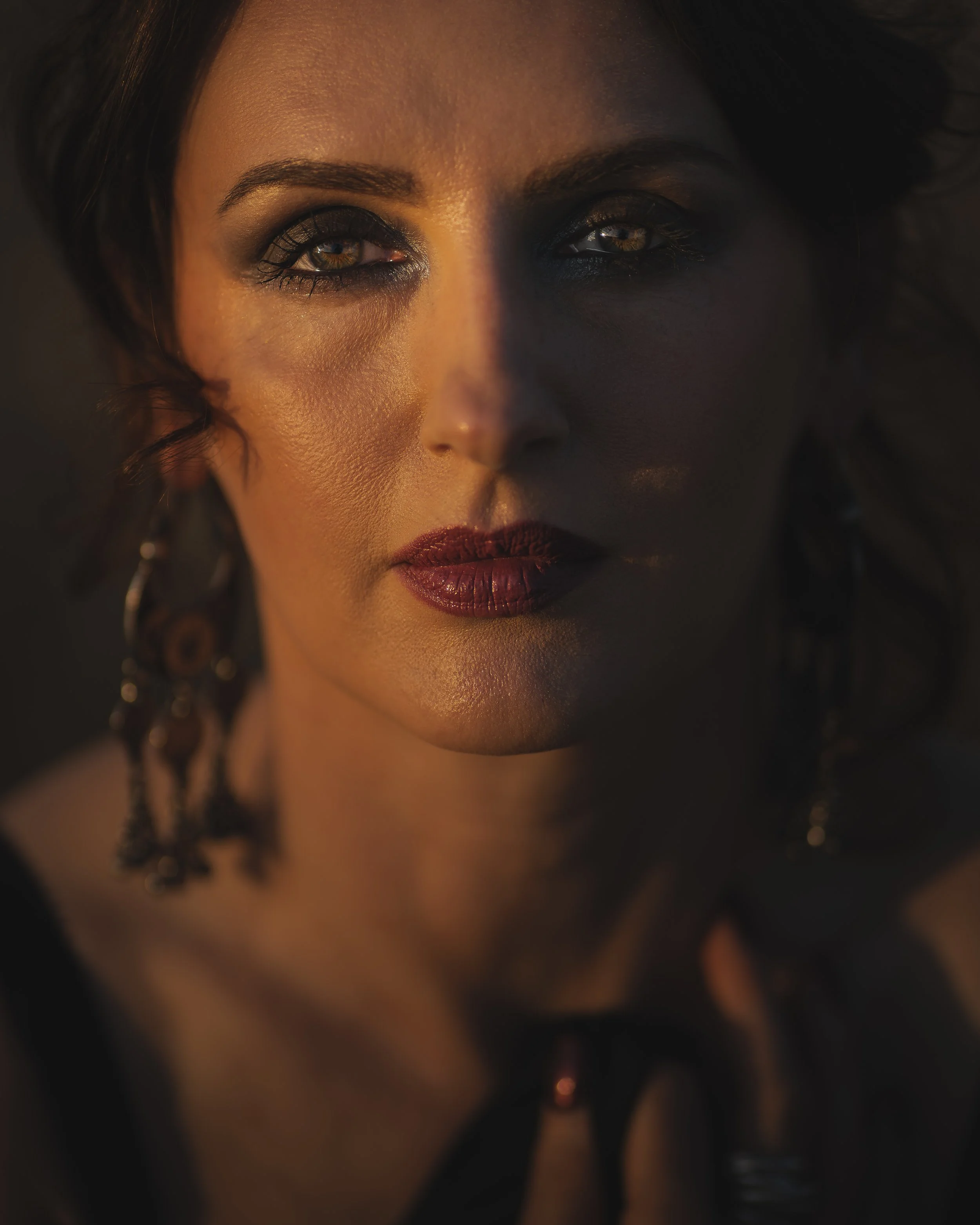 Close-up of a woman's face with dramatic makeup, dark eyeshadow, and deep red lipstick, wearing earrings and a dark top, in dim lighting.
