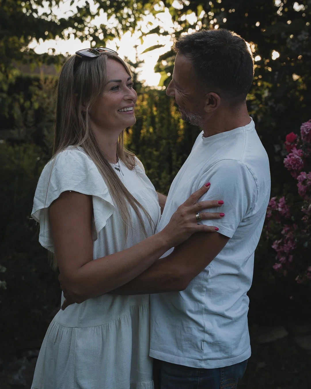 A man and woman smiling and looking at each other while holding each other in a garden during sunset.