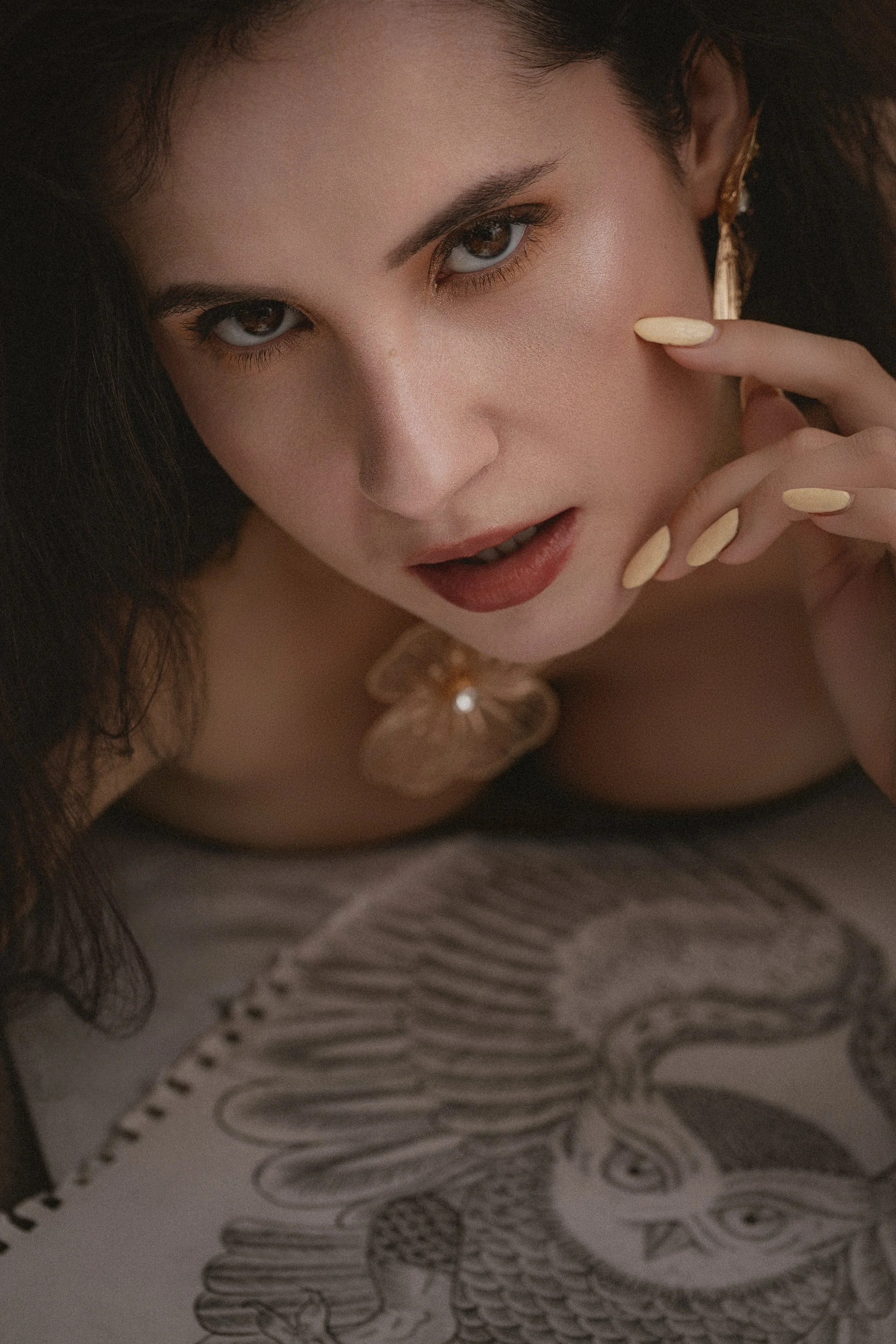 Close-up portrait of a woman with dark curly hair, brown eyes, and light makeup, resting her chin on her fingers, with a drawing in the background.