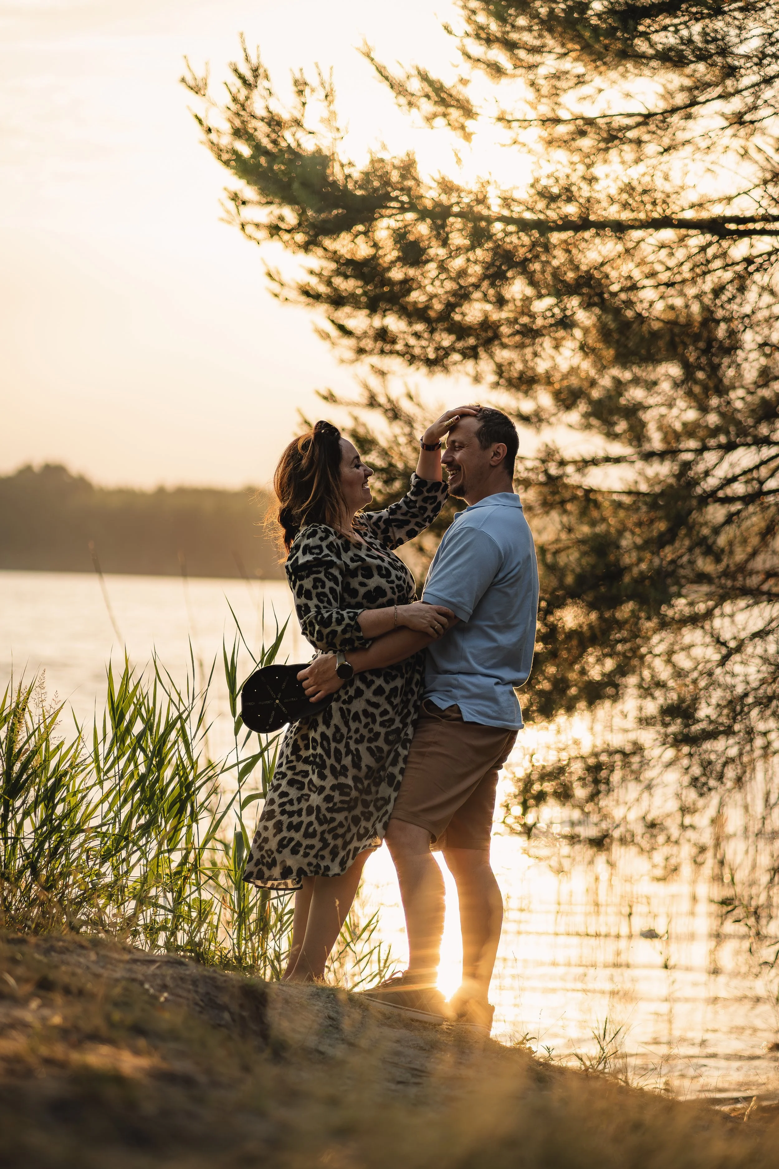A couple standing near a lake during sunset, smiling and touching foreheads, with trees in the background.