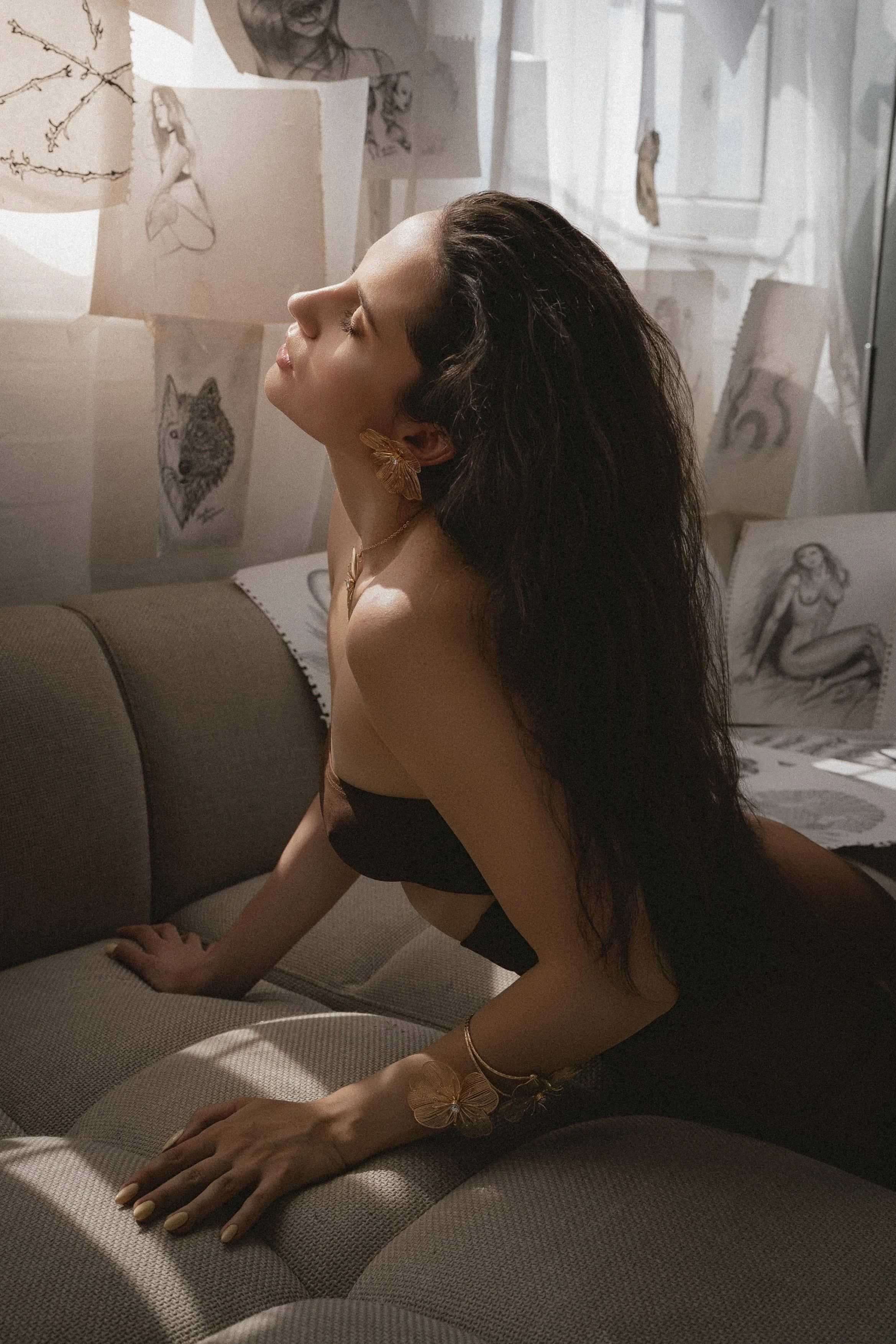 A woman with long dark hair, wearing a butterfly-shaped earring and a bracelet, is posed on a beige couch with her eyes closed and head tilted back. She is in a room with sketches and drawings of women and animals taped to the wall behind her.