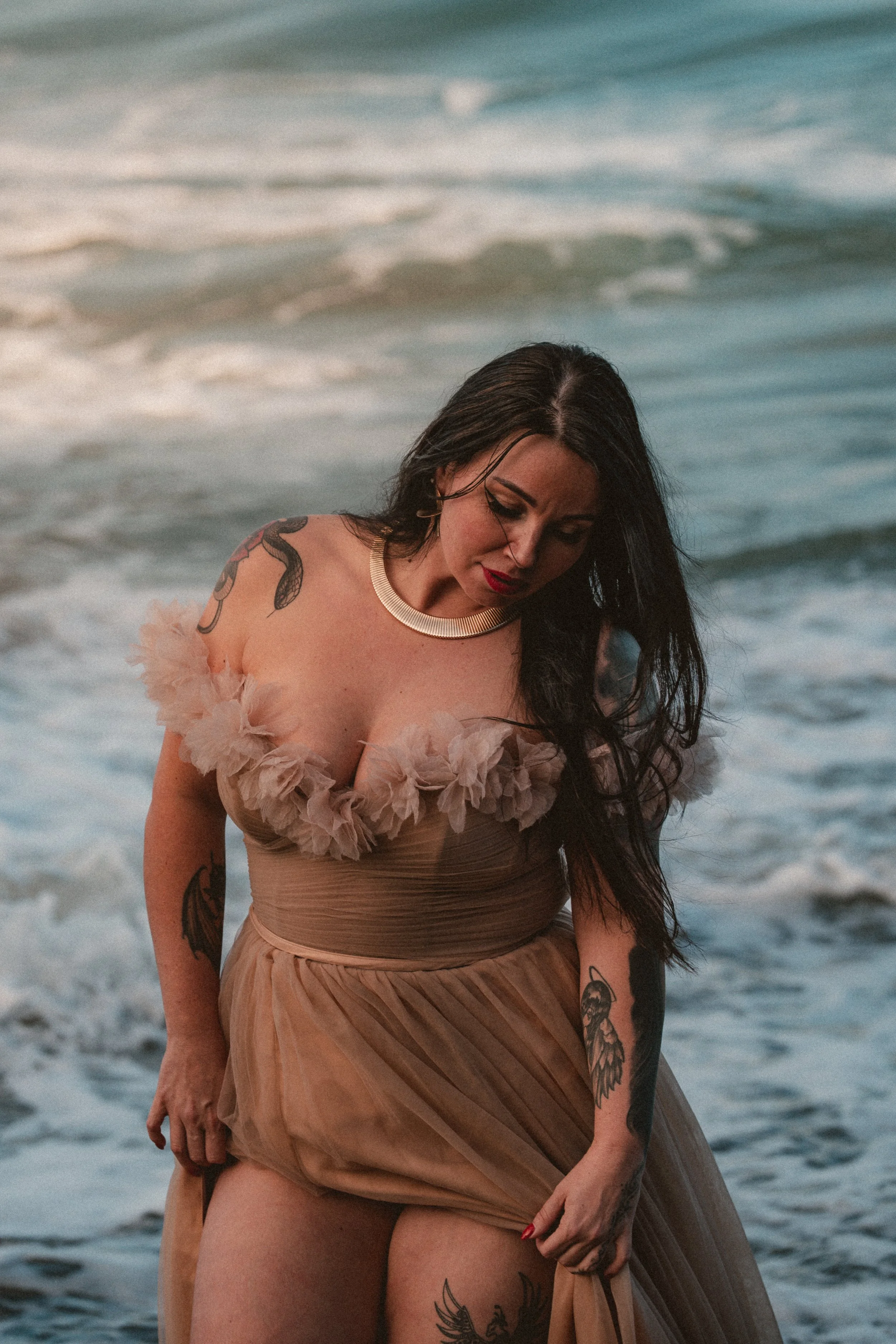 Woman with dark hair and tattoos wearing a beige dress with floral details at the beach, standing in front of the ocean waves.