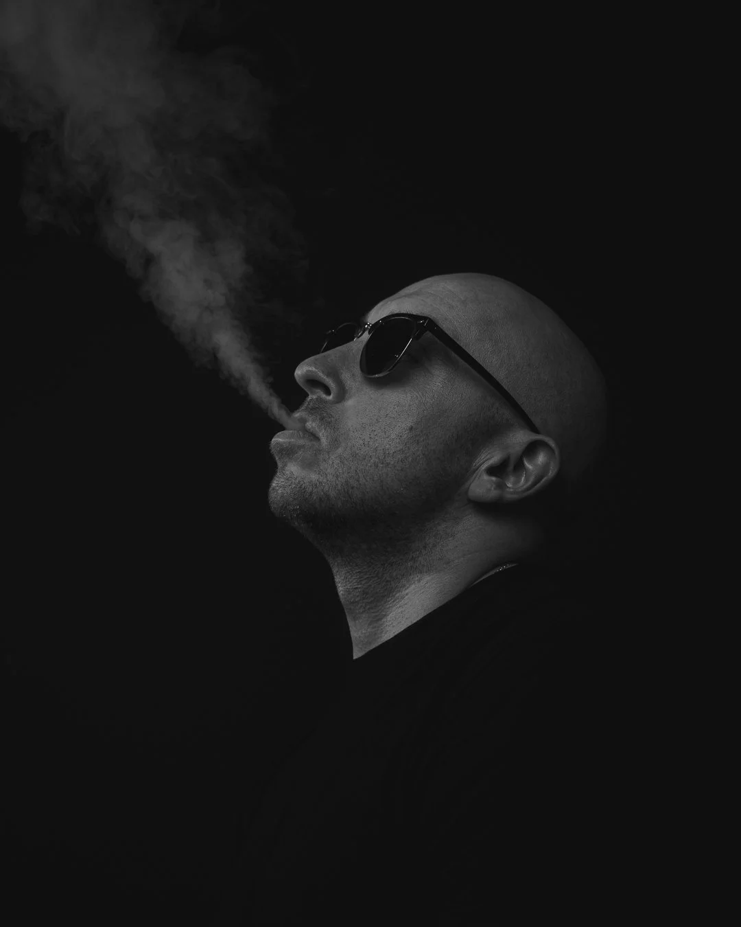 A black-and-white photo of a man wearing sunglasses and a head covering, smoking with smoke coming out of his mouth.