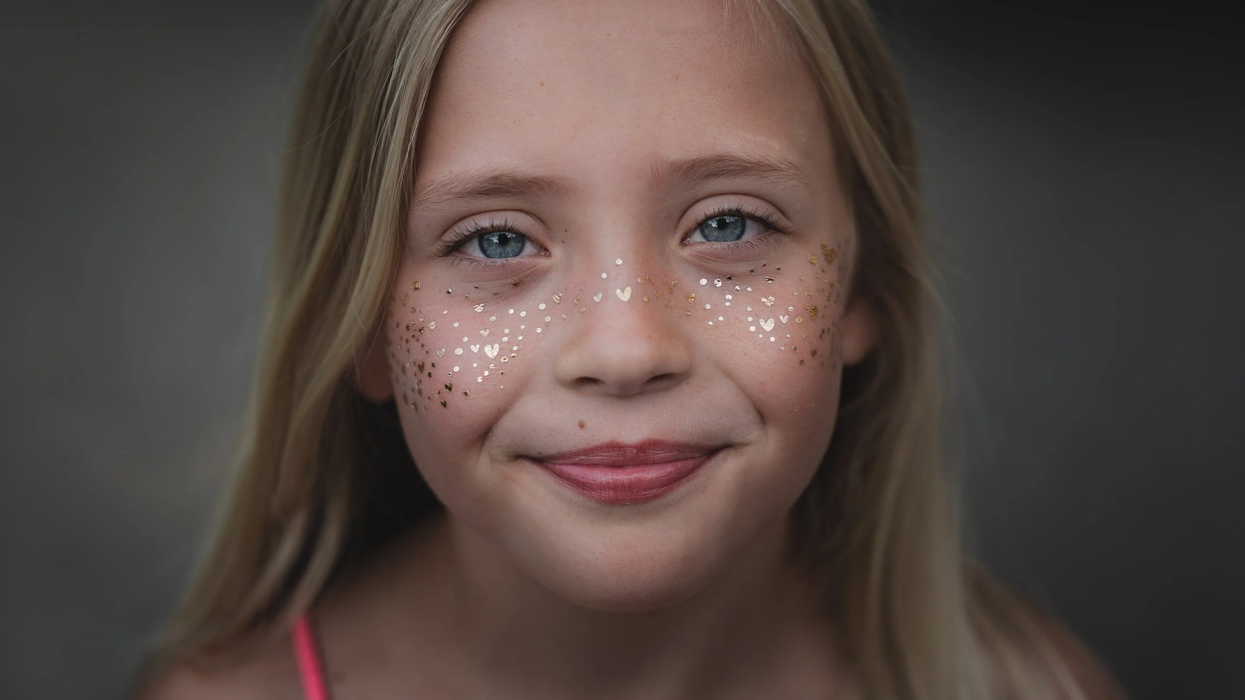 Close-up of a young girl with light blonde hair, blue eyes, and gold heart-shaped face glitter, smiling.