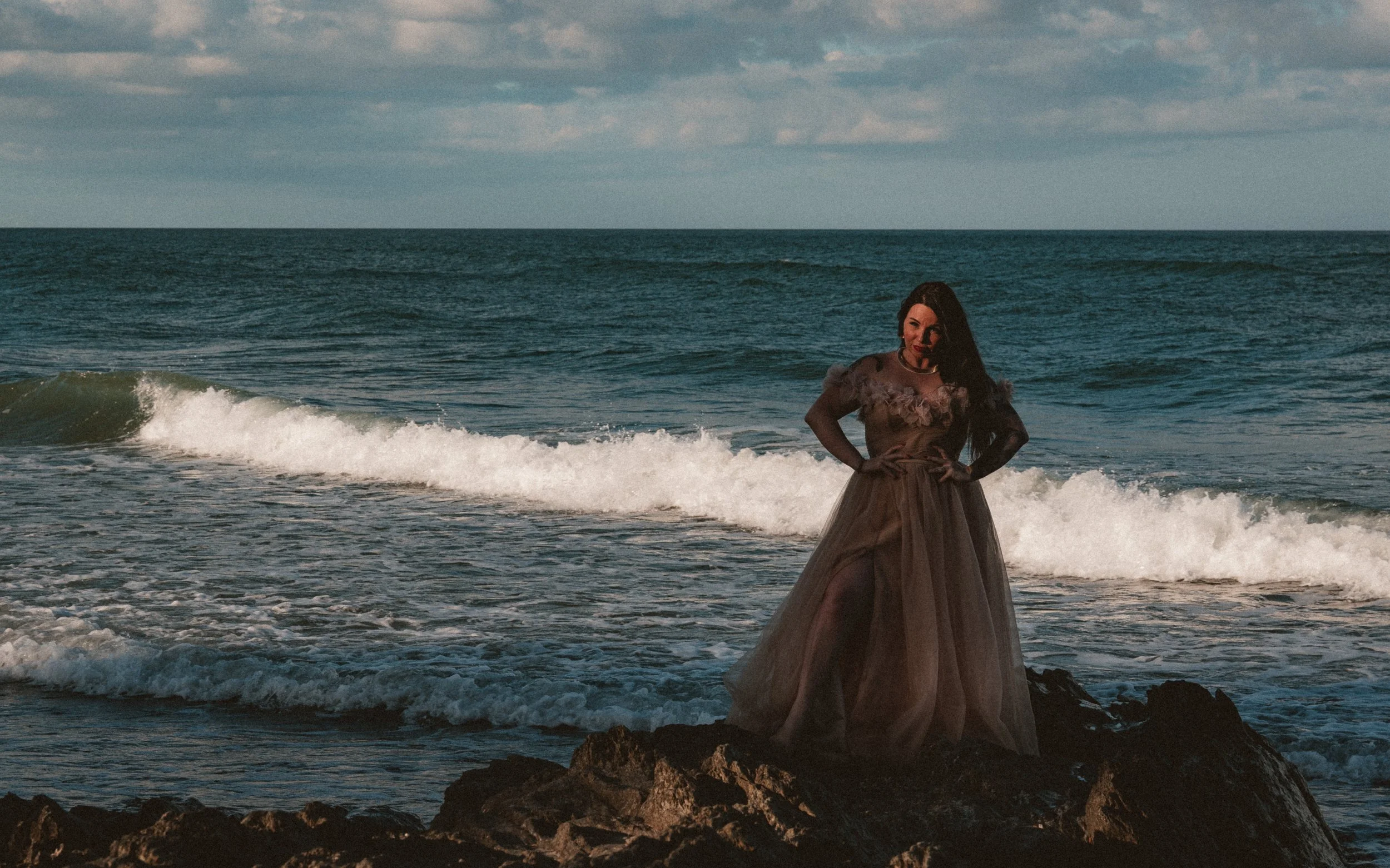 A woman in a formal gown standing on rocks at the beach with the ocean waves in the background during sunset or dusk.