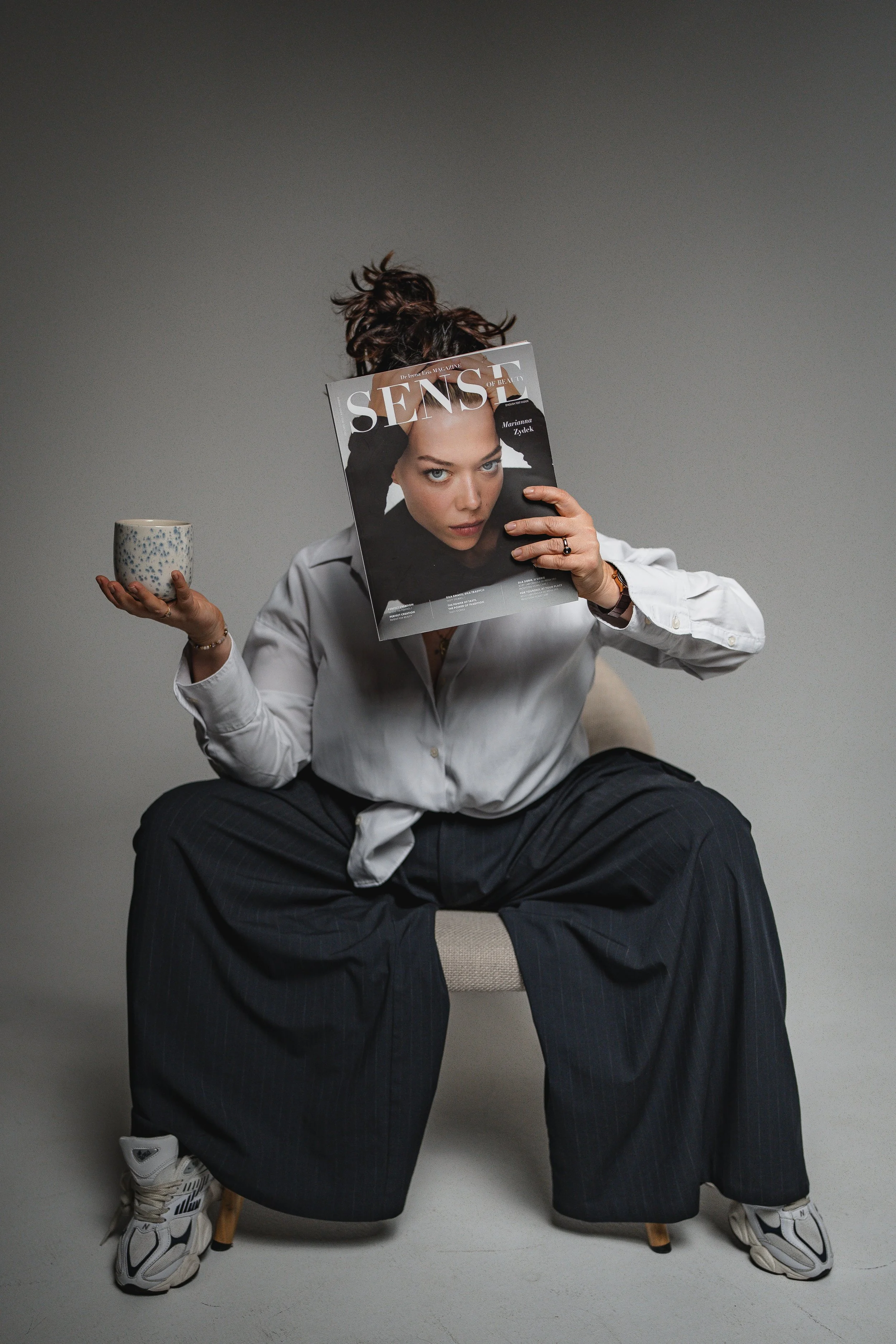 Person sitting on a chair, holding a cup in one hand and a magazine in front of their face. The person is dressed in a white shirt and dark striped pants, wearing sneakers, with their legs spread apart and feet flat on the ground.