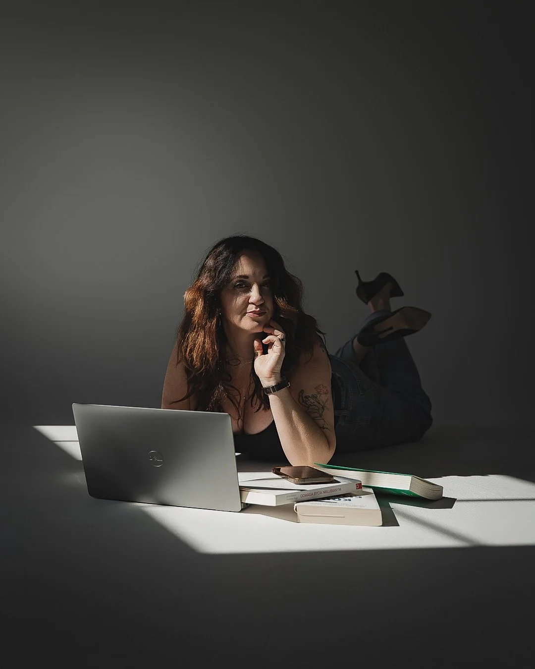 Woman lying on her stomach at a desk with a laptop, books, a notebook, and a smartphone, illuminated by a focused light with a dark background.