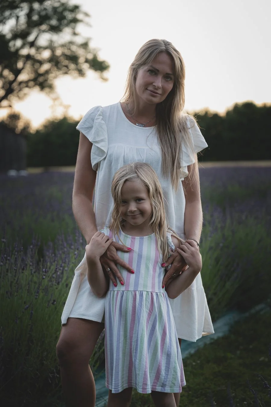 A woman and a young girl standing in a lavender field during sunset, smiling at the camera.