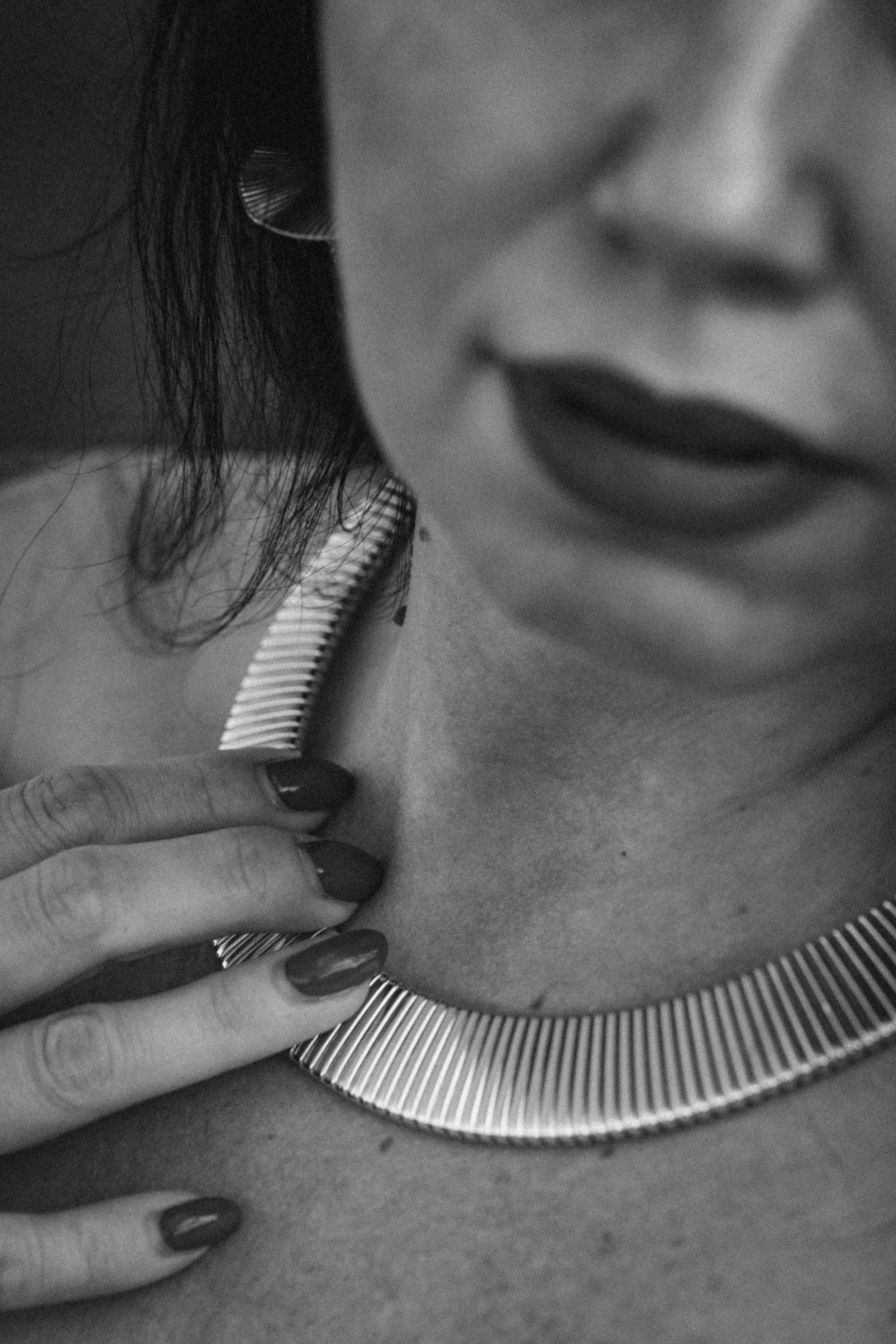 Close-up black and white photo of a person touching their neck with their fingers, with sharp painted nails, next to a round mirror with a textured edge, showing part of their face and lips.