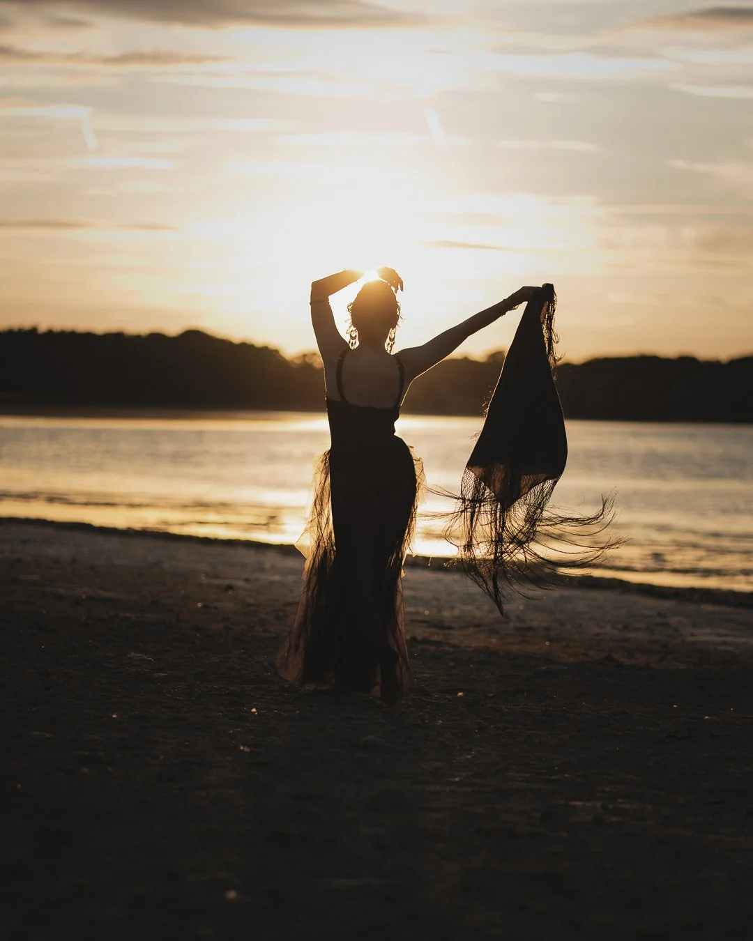Silhouette of a woman on a beach at sunset, stretching and holding a piece of cloth, with water and trees in the background.