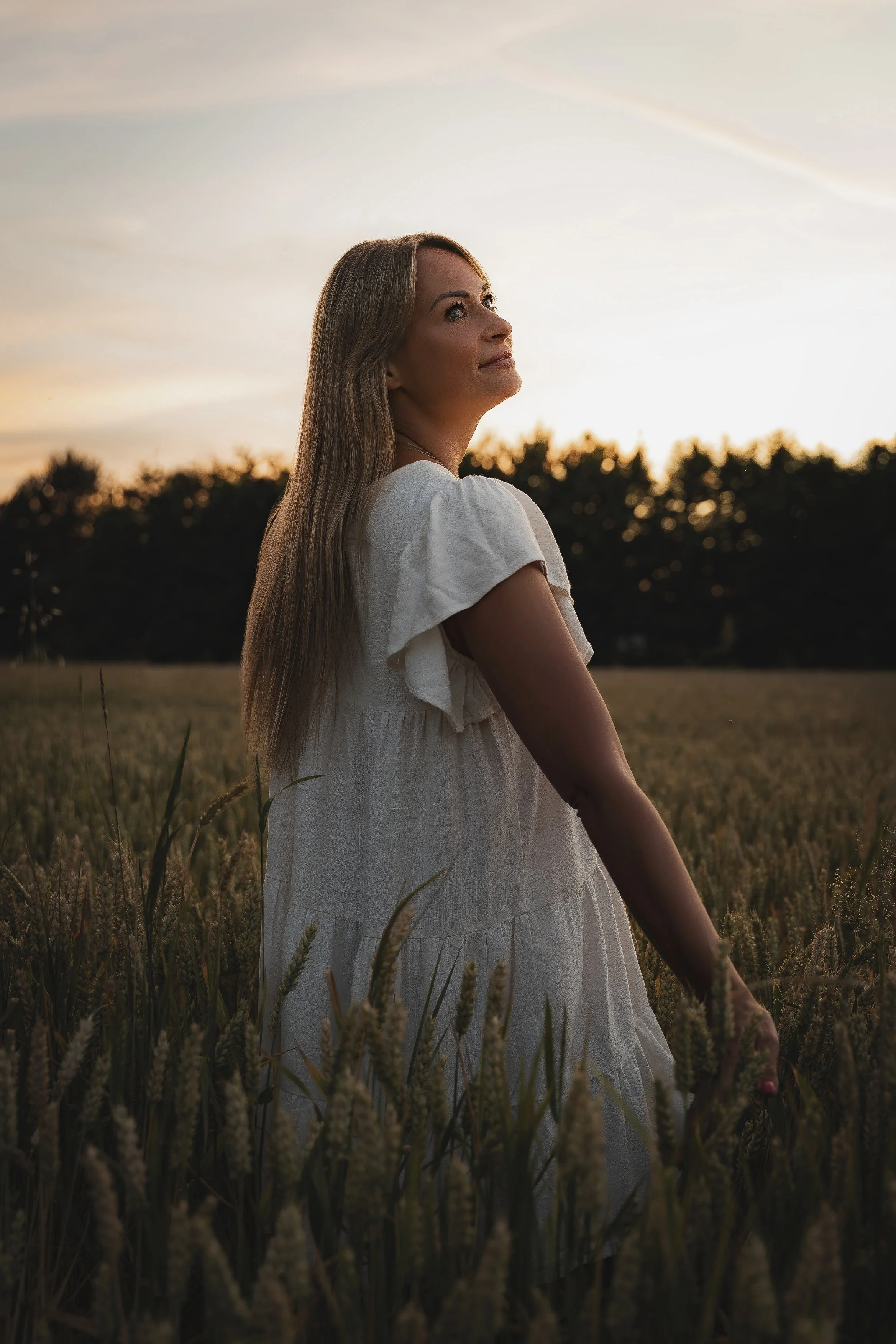 A woman in a white dress standing in a wheat field at sunset, looking upward with a peaceful expression.