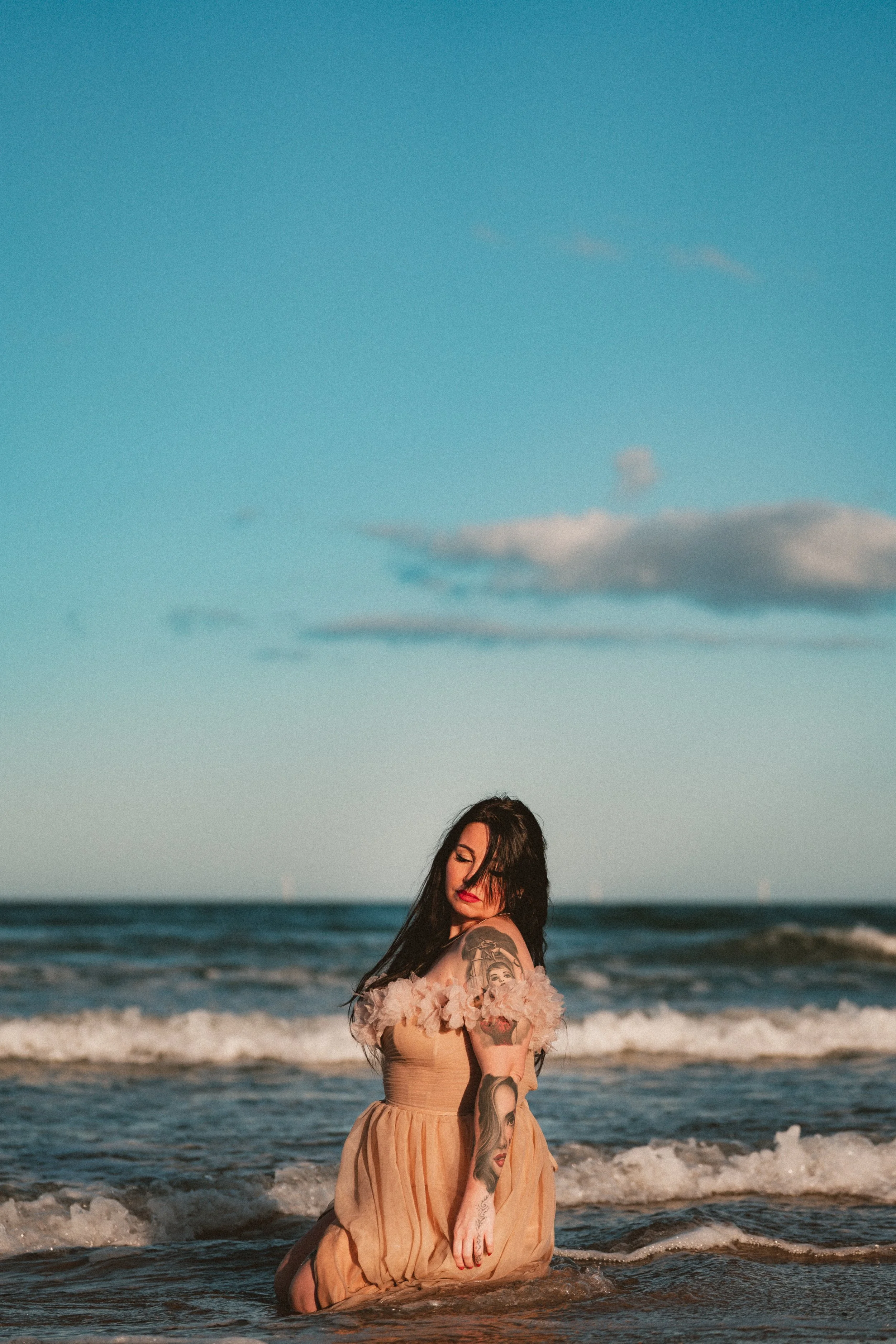 A woman with dark hair and tattoos on her arm, wearing a beige dress with fluffy pink sleeves, sits in the ocean with waves around her and a blue sky with clouds in the background.