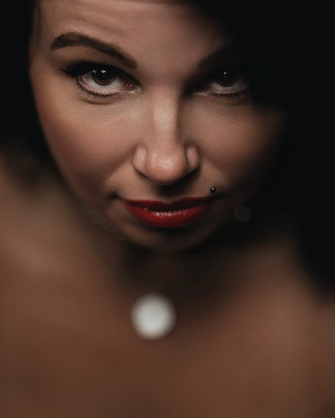 Close-up of a woman's face with dark hair, makeup, and a nose piercing, looking directly at the camera.