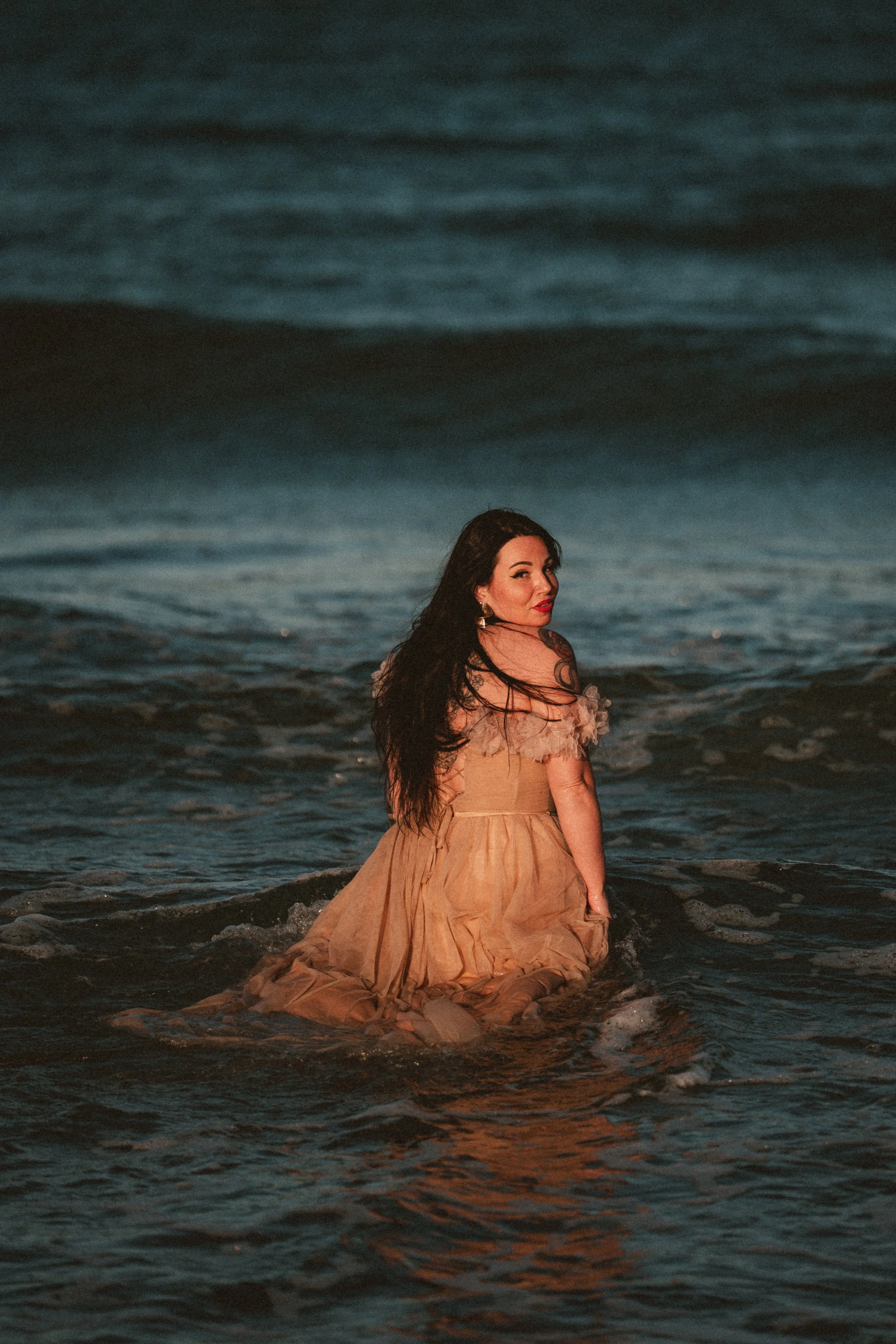 A woman with long dark hair wearing a light-colored dress sitting in the shallow water at the beach during dusk, with waves around her and a dark sky in the background.