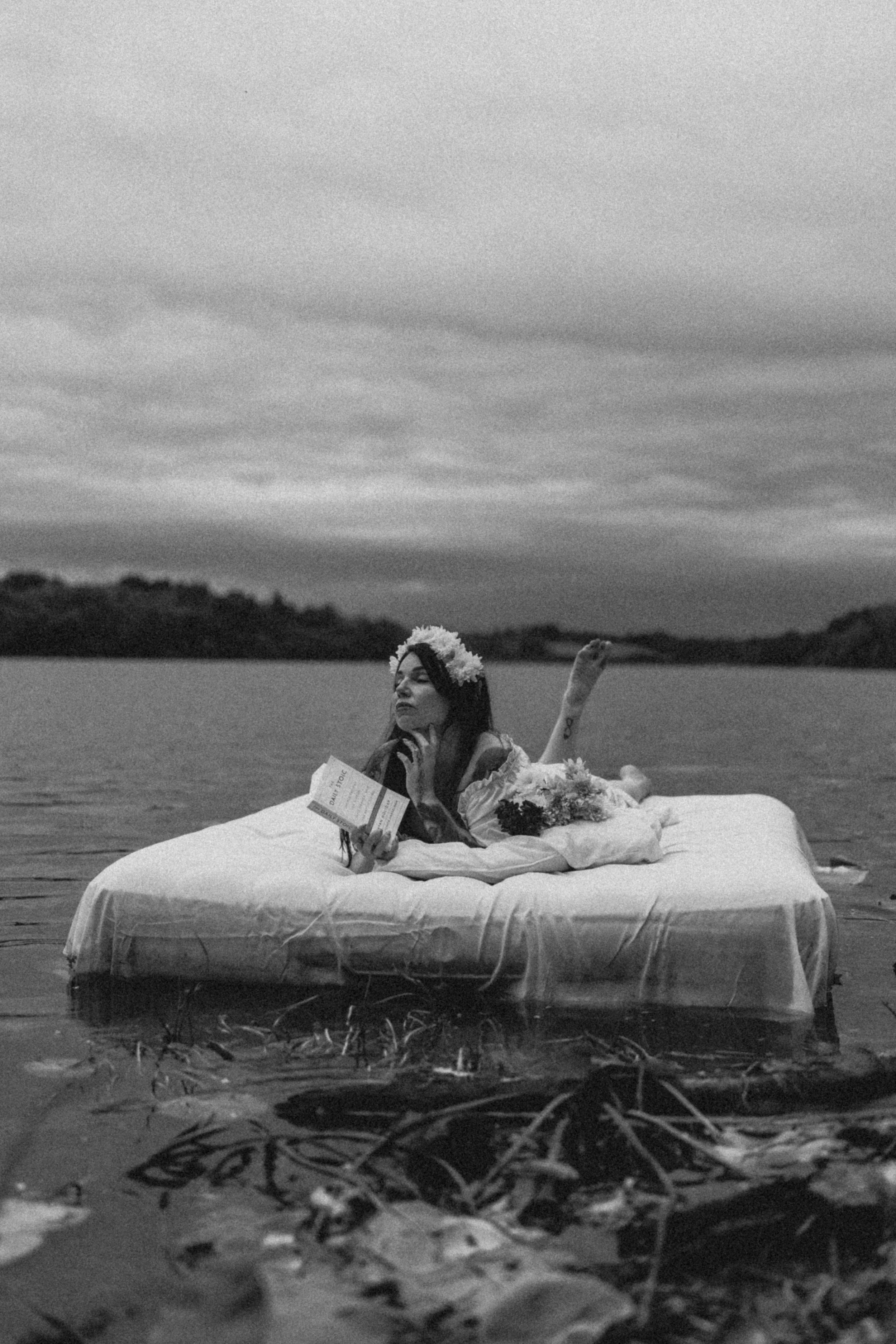 A woman lying on a large bed on a body of water, holding a book, with a flower crown on her head, and surrounded by flowers, with cloudy skies in the background.