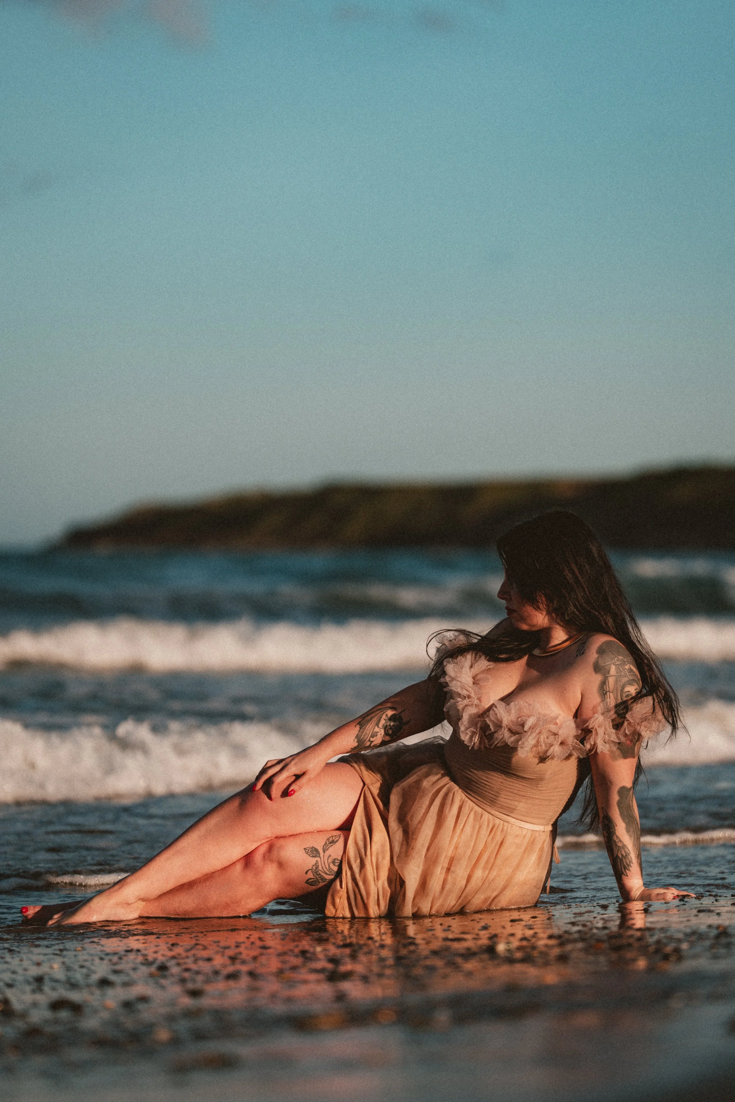 A woman with tattoos on her arms and legs, sitting on the beach with her legs extended in front of her and her left hand resting on her knee. She is wearing a beige dress with tulle and ruffles and has dark hair. The ocean waves are in the background, and the sky is clear.