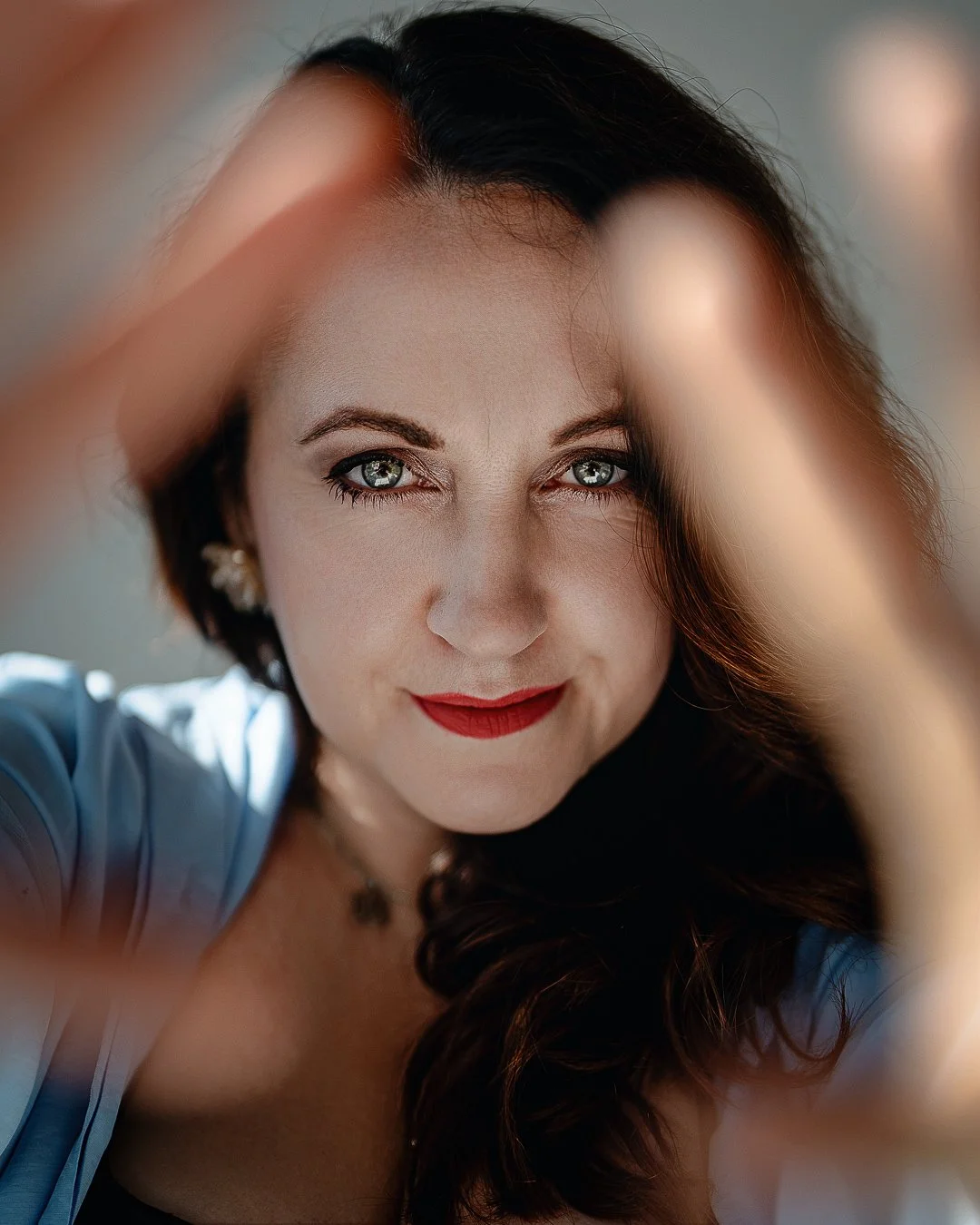 A woman with dark hair and gray eyes looking directly at the camera, her face framed by blurred hands in the foreground, wearing red lipstick and earrings.