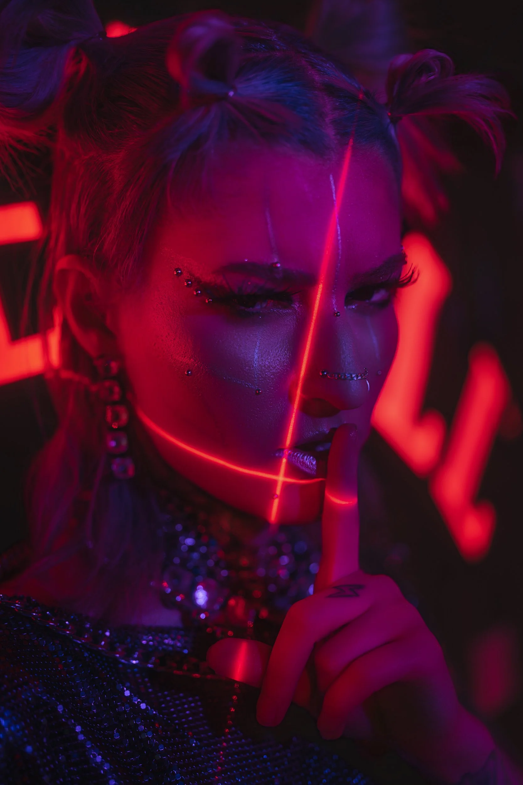A woman with light hair styled in two buns, wearing eye makeup and face jewelry, holds her finger to her lips in a shushing gesture. She is illuminated by neon red and purple lights, creating a futuristic and edgy atmosphere.