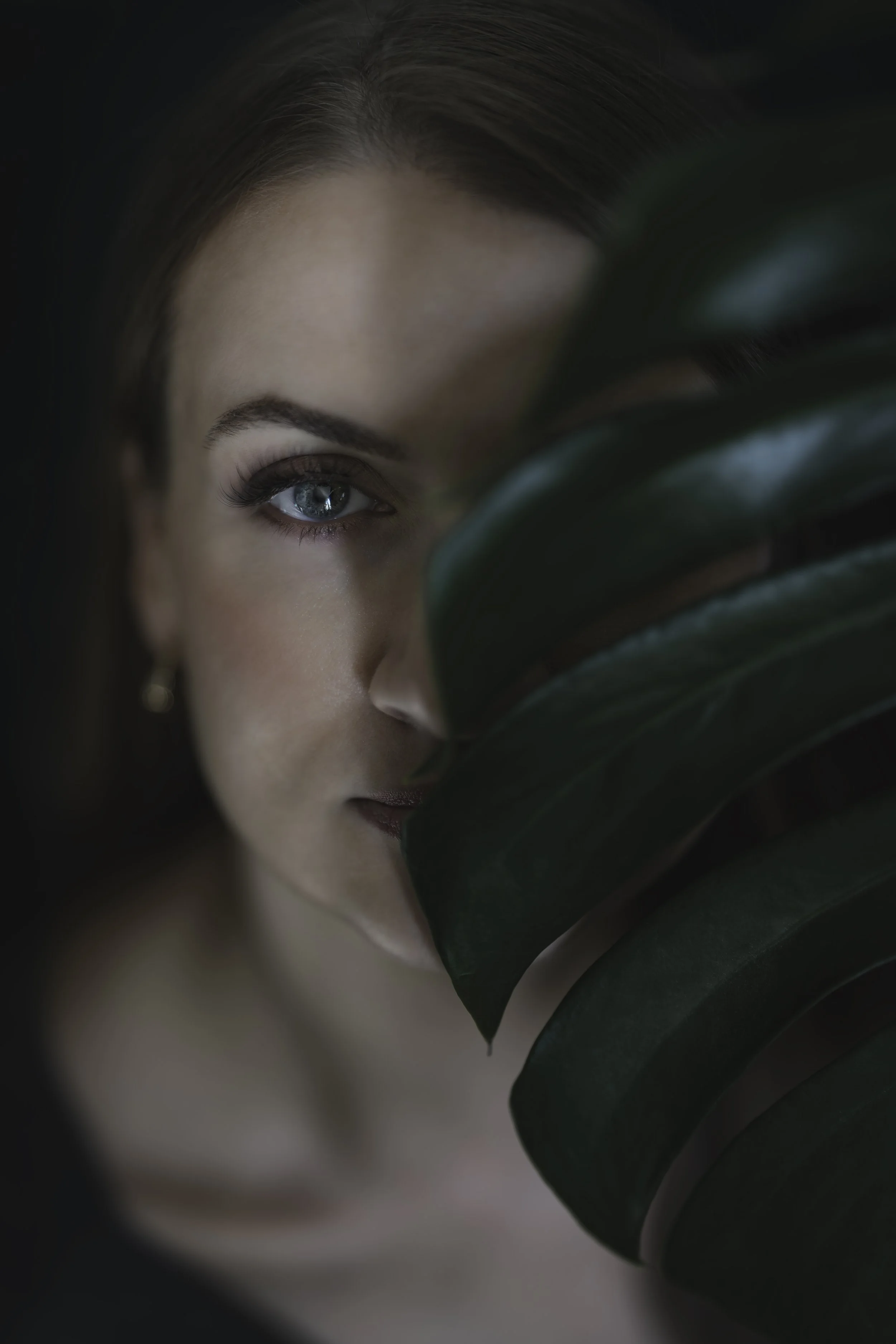 Close-up of a woman with blue eyes partially obscured by large green leaves, featuring natural makeup and dark hair.