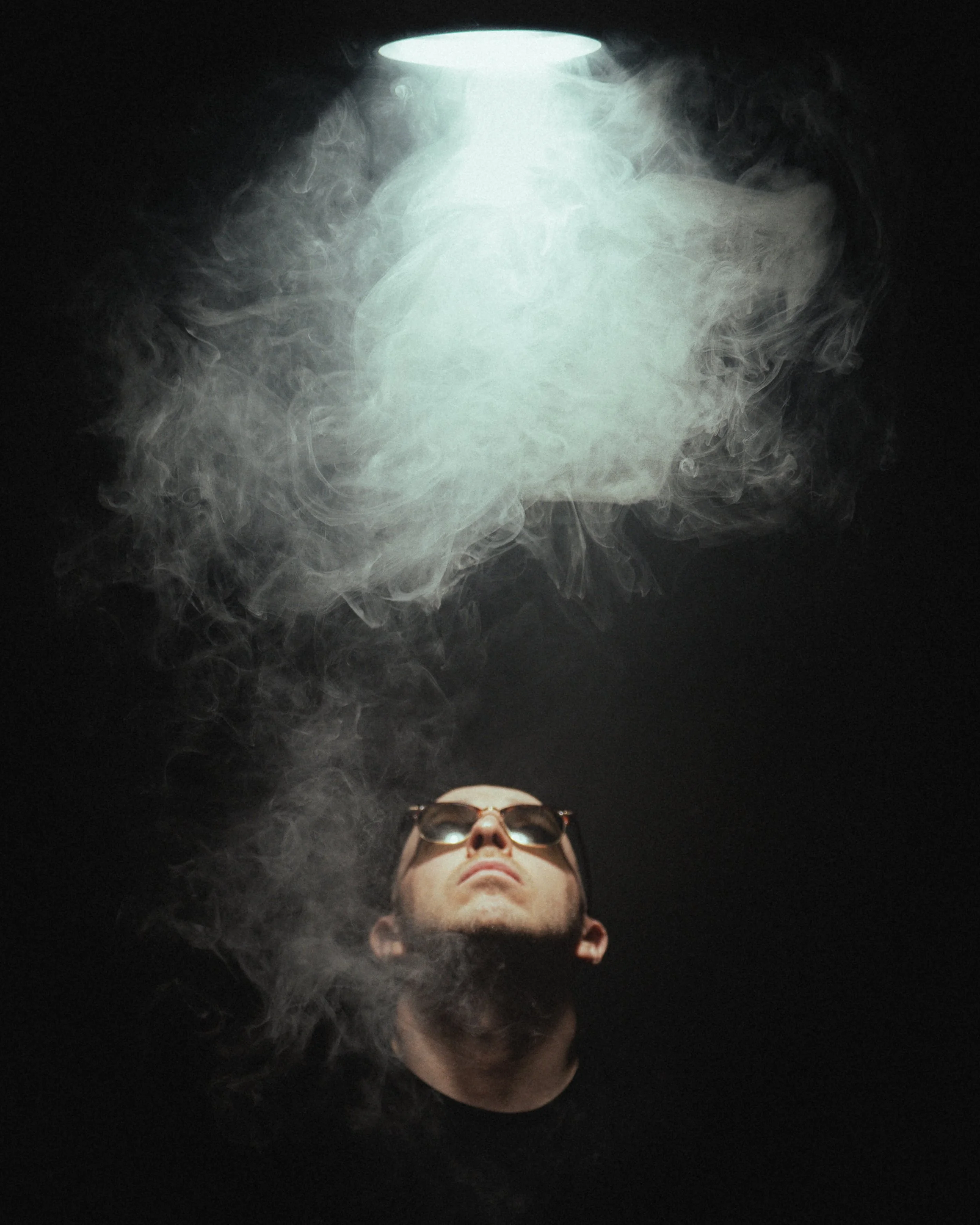A man wearing sunglasses looking up at a light source with smoke around him in a dark room.