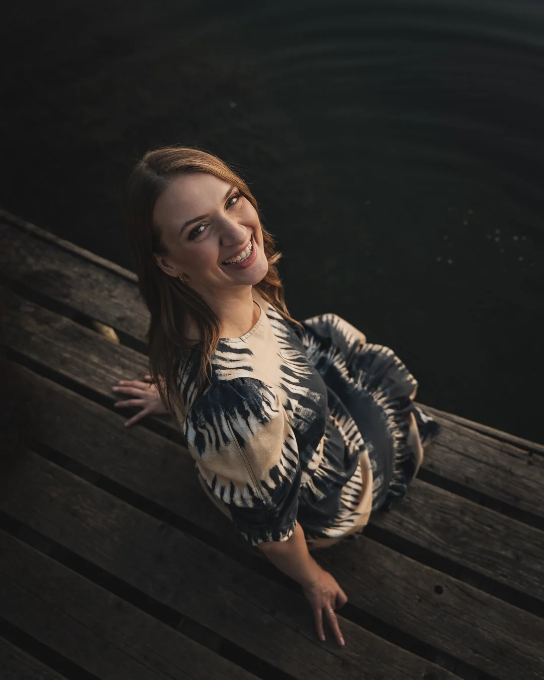 A woman sitting on a wooden dock by the water, smiling and looking up at the camera.