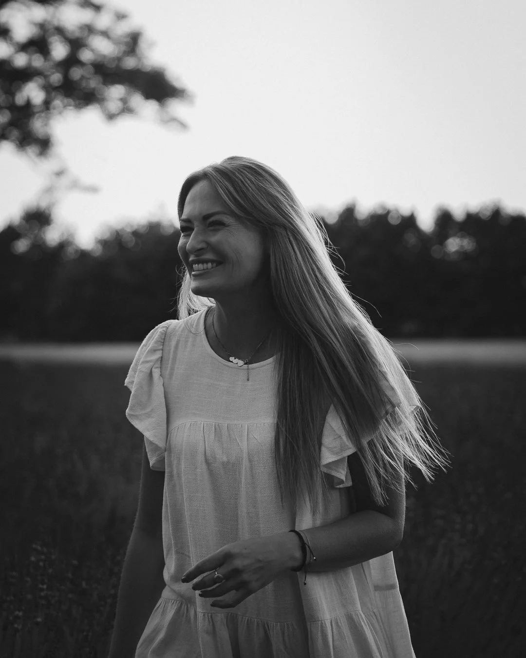A smiling woman with long blonde hair wearing a light-colored dress standing outdoors in a field with trees in the background, captured in black and white.