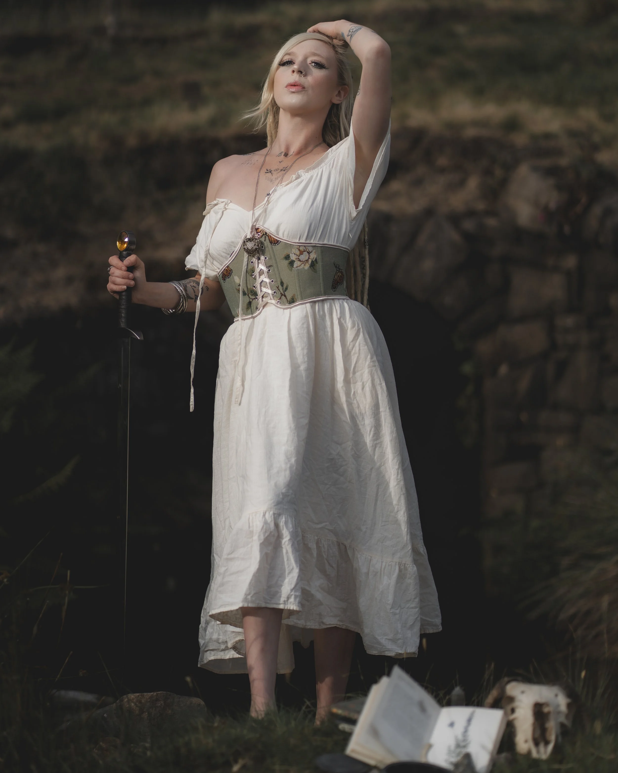 A woman standing outdoors near a rock archway, dressed in a white dress with a floral corset, holding a sword in her right hand, with a book and skull at her feet.