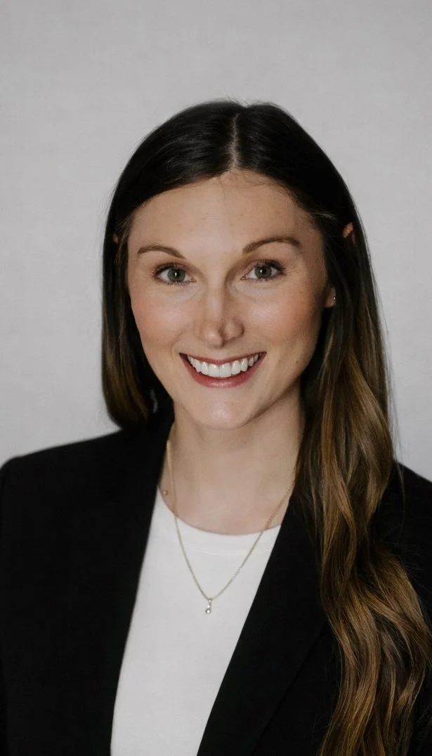 A young woman with long brown hair wearing a black blazer, a white shirt, and a delicate necklace, smiling at the camera against a plain light-colored background.