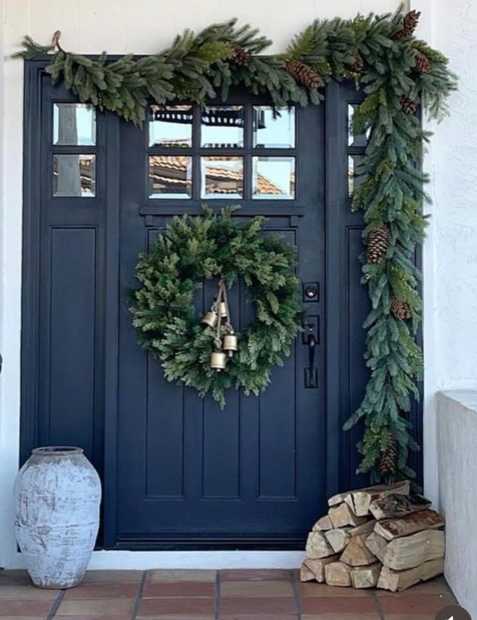 A dark blue front door decorated with a green wreath hanging in the center and a garland of evergreen branches with pinecones adorning the sides and top. There is a white, rustic vase on the left and a pile of chopped firewood on the right, on a tile