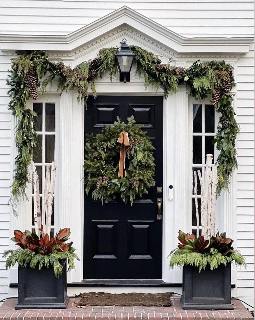 Front door decorated with a Christmas wreath, greenery garland with pinecones, and potted plants on either side.