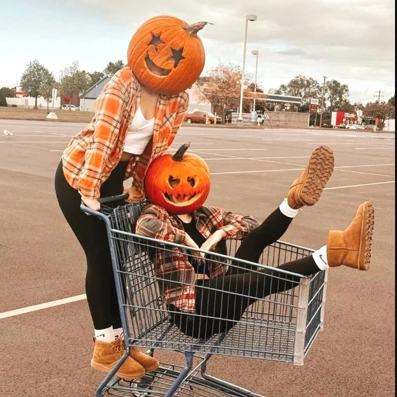 Two people with pumpkin masks on their heads, one sitting inside a shopping cart and the other pushing, both wearing plaid shirts and boots, in an empty parking lot.