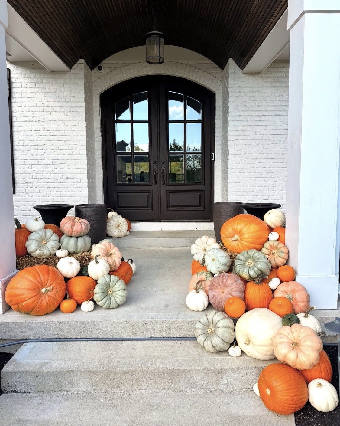 Front porch decorated with a large collection of pumpkins in various colors, including orange, white, green, and pink, arranged on the steps and sides of the porch leading to a black door with glass panels.