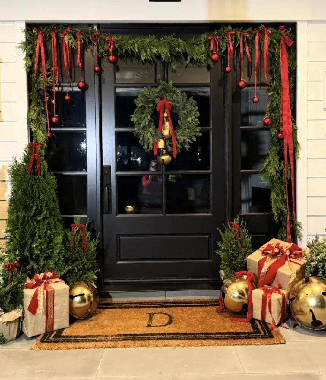 Festively decorated front porch with a black door, green greenery garland with red ribbons and ornaments, a wreath with candles, and wrapped gifts with red ribbons, accompanied by gold bells and potted plants.