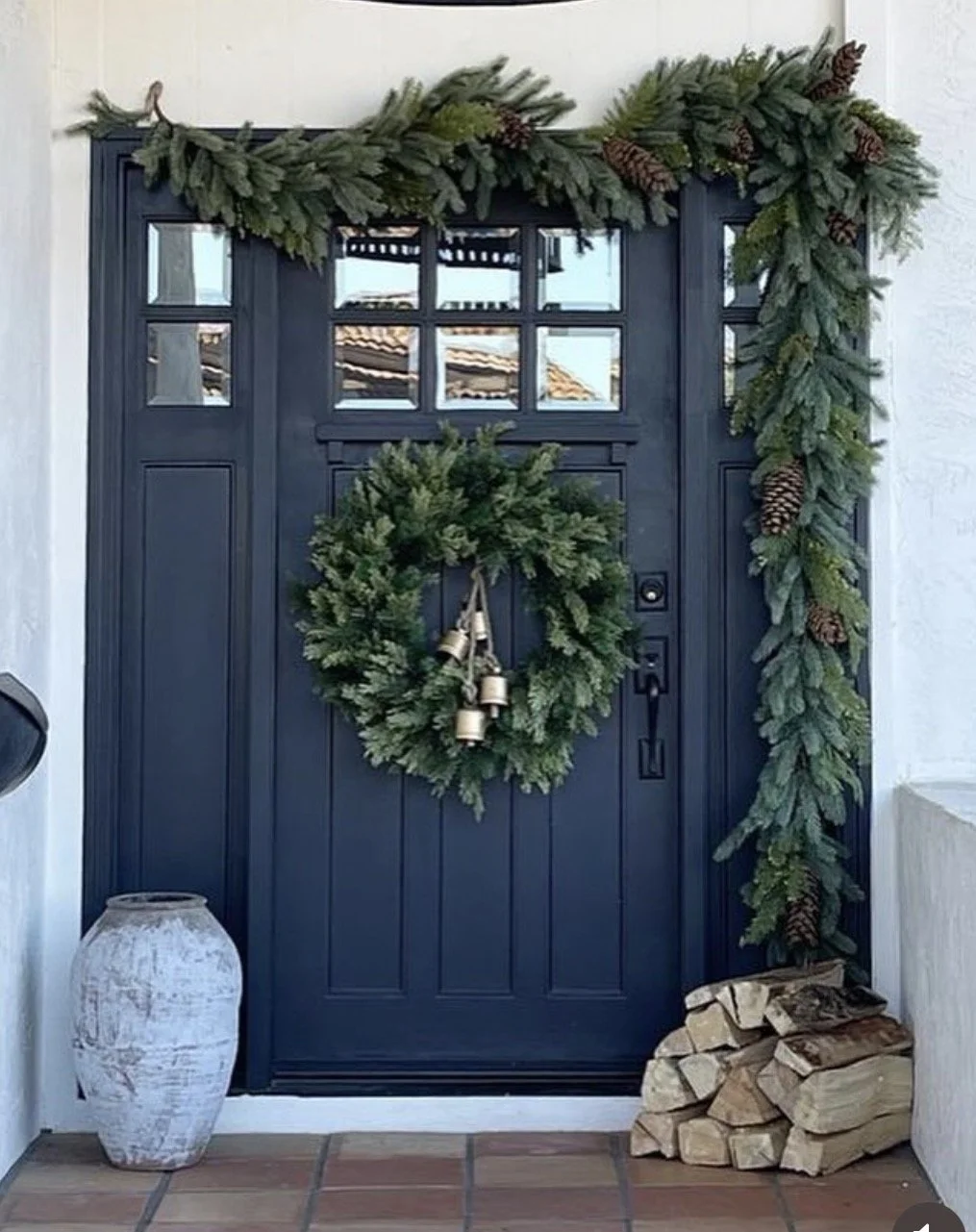 A dark blue front door decorated with a green wreath hanging in the center and a garland of evergreen branches with pinecones adorning the sides and top. There is a white, rustic vase on the left and a pile of chopped firewood on the right, on a tile