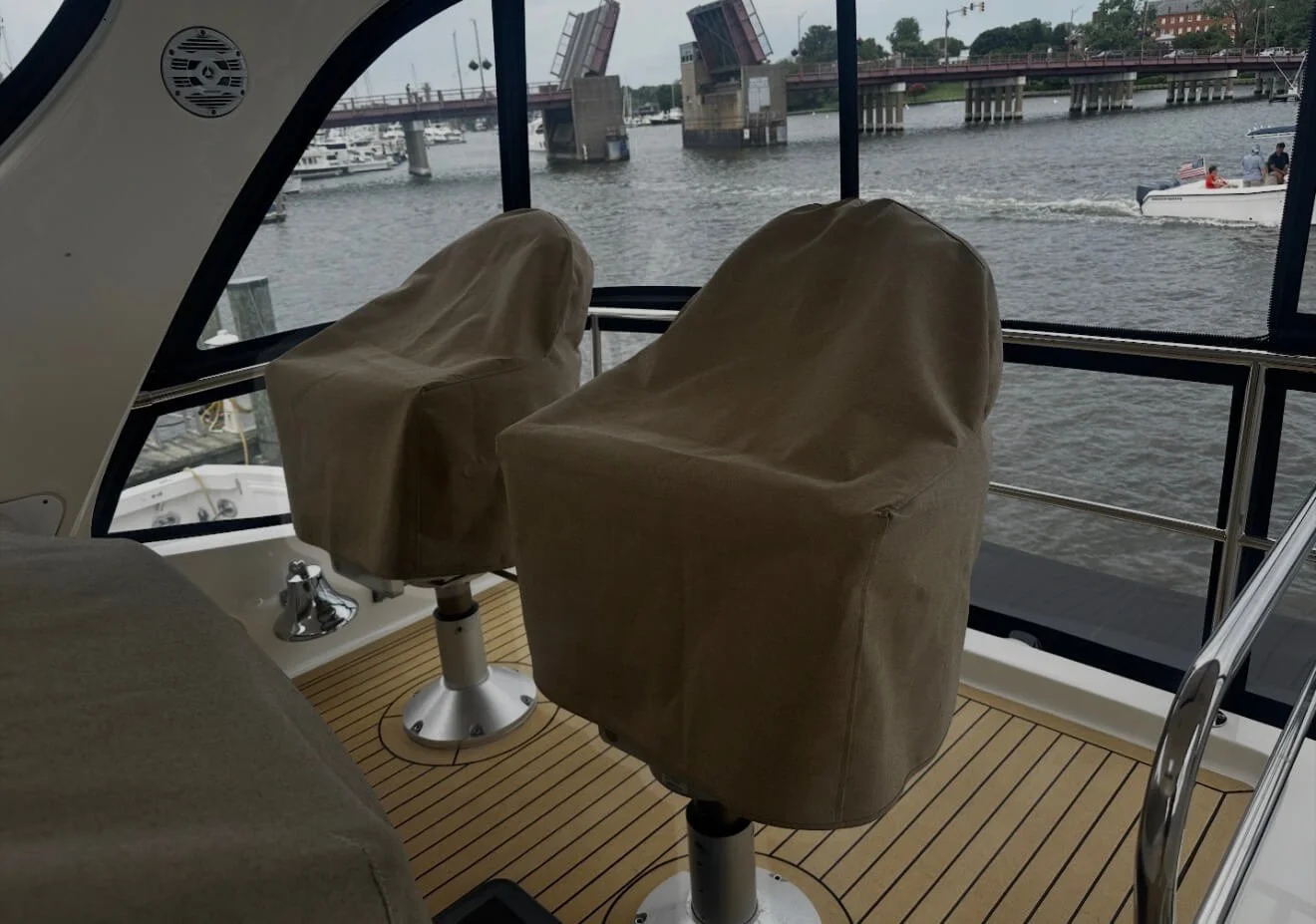 Two captain's chairs covered with beige fabric inside a boat with a view of water, a bridge, and boats outside.