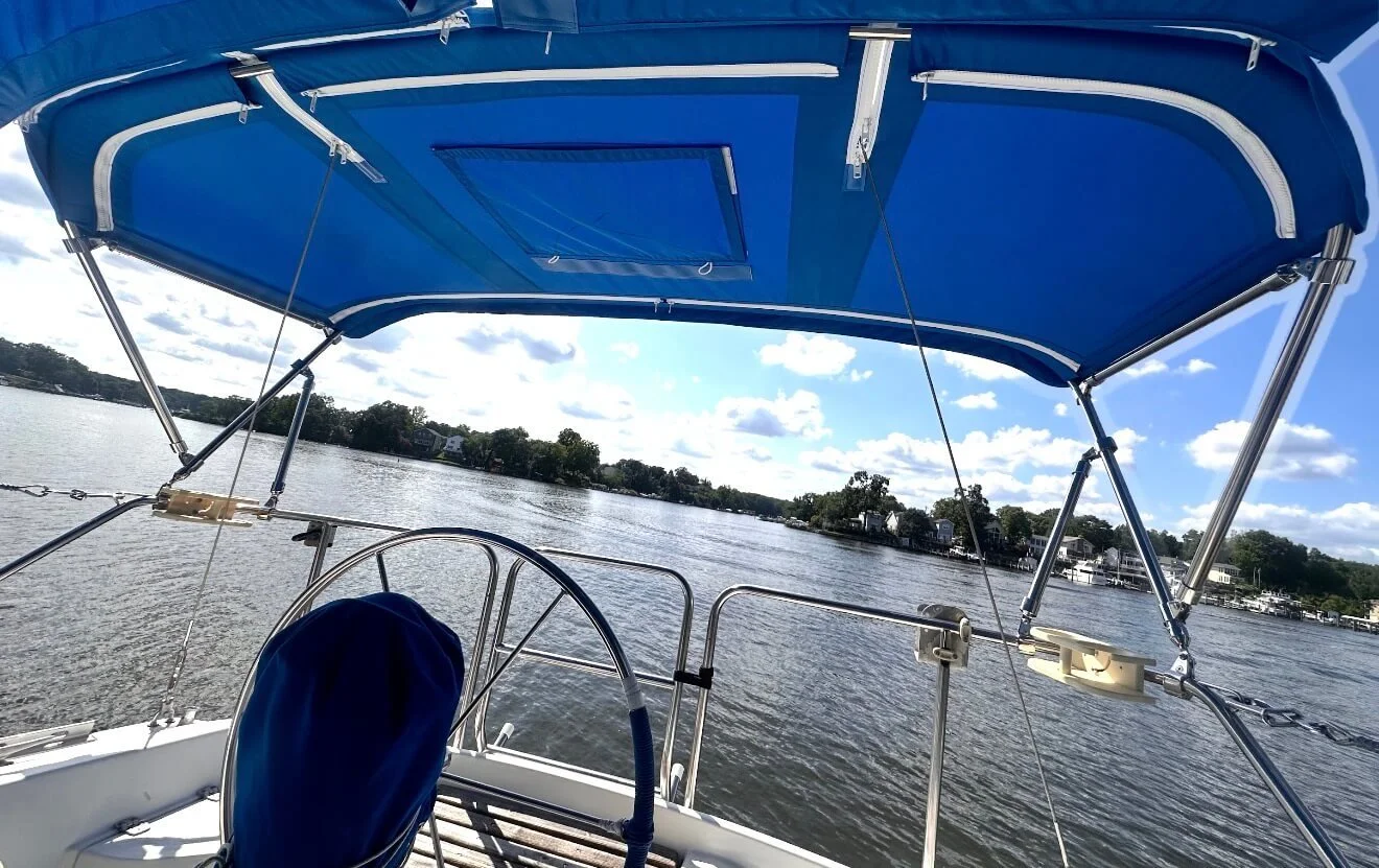 View from a boat on a river, showing the boat's blue canopy, steering wheel, and a blue seat cover. On the river, there is water with some houses and trees along the shoreline under a partly cloudy sky.