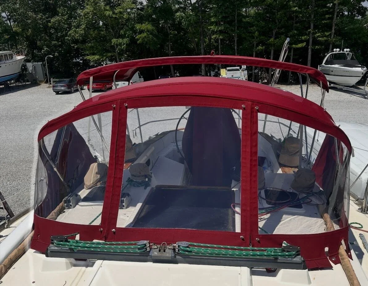 Front view of a sailboat with a red canopy and clear plastic windows, docked at a boat yard with trees and other boats in the background.