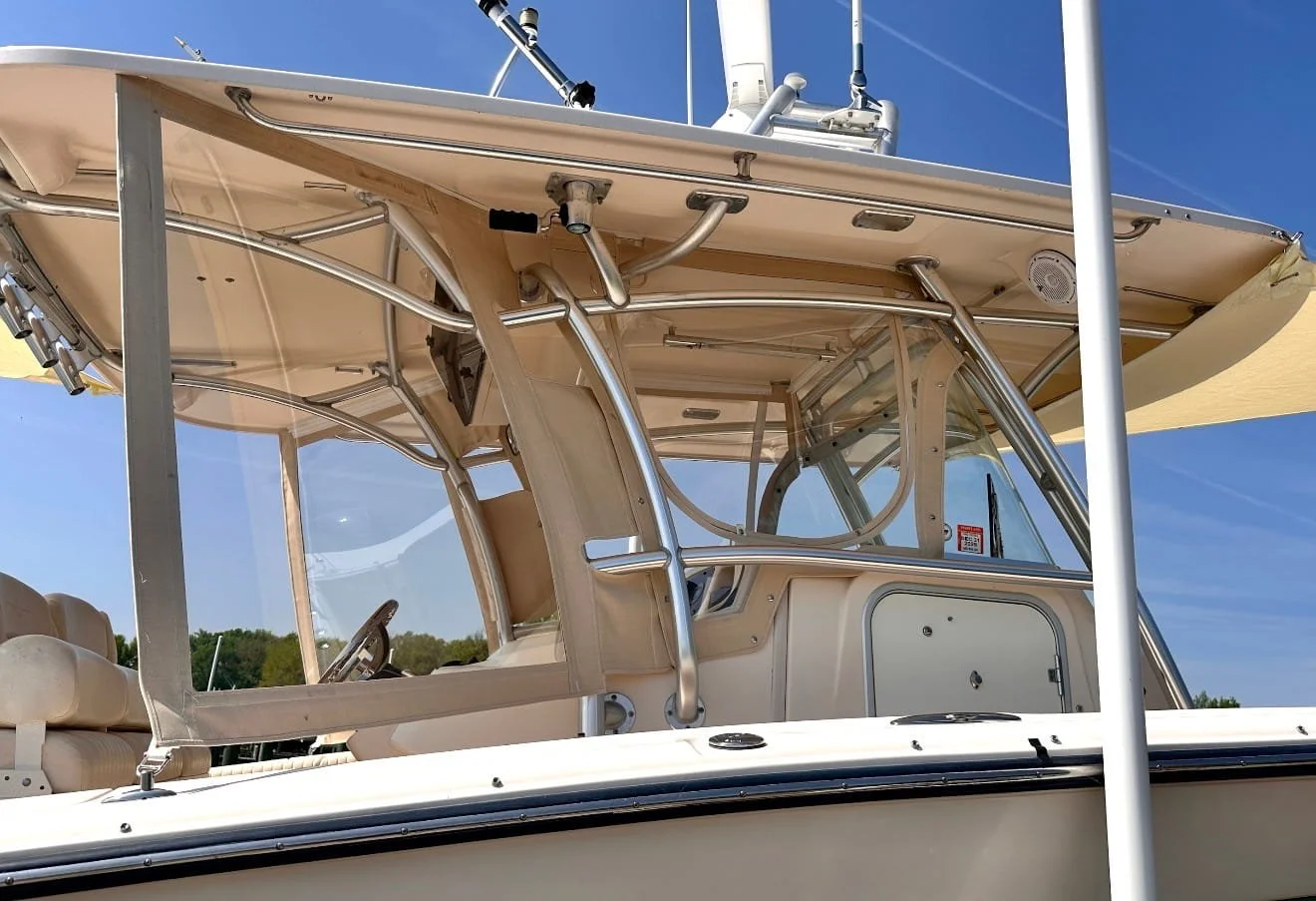 A close-up of a boat's cockpit with beige seating, metal railings, and a canopy, against a blue sky and some greenery in the background.