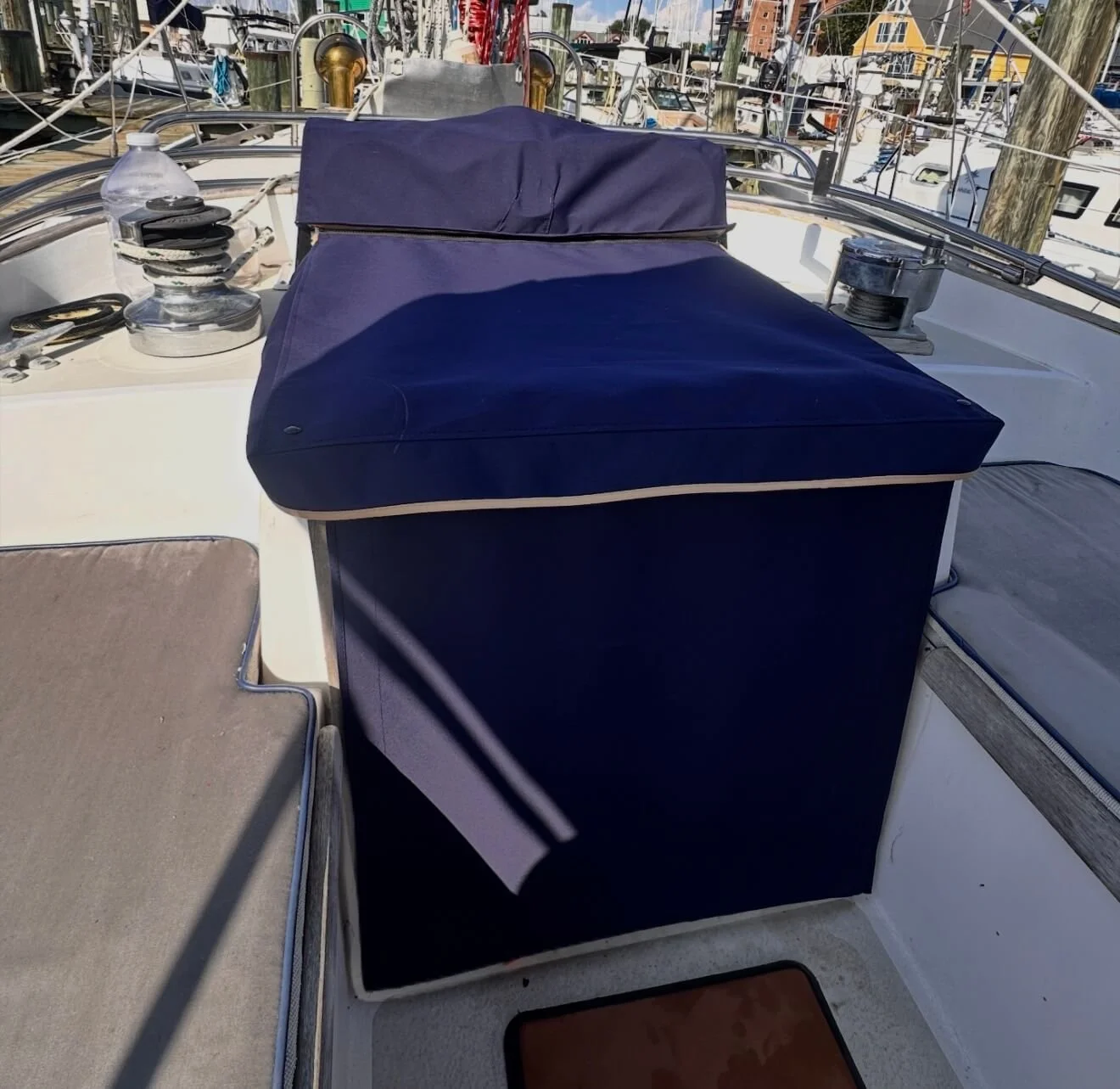 A blue and white yacht deck with a blue cushioned seat and various equipment, including a water bottle and winches, docked at a marina with other boats in the background.