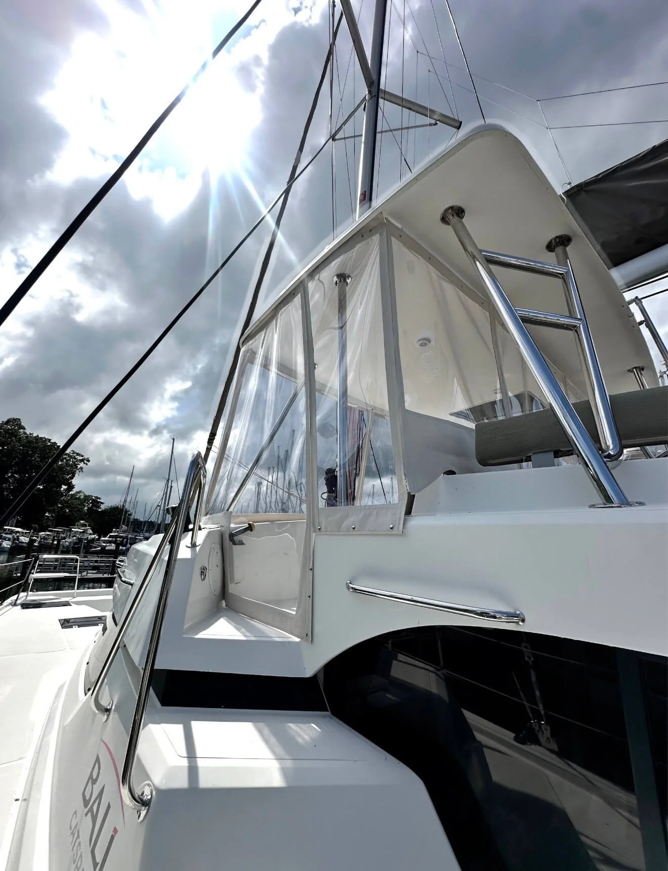 View of a white yacht docked at a marina, with rigging and masts visible, partly cloudy sky, and sunlight.