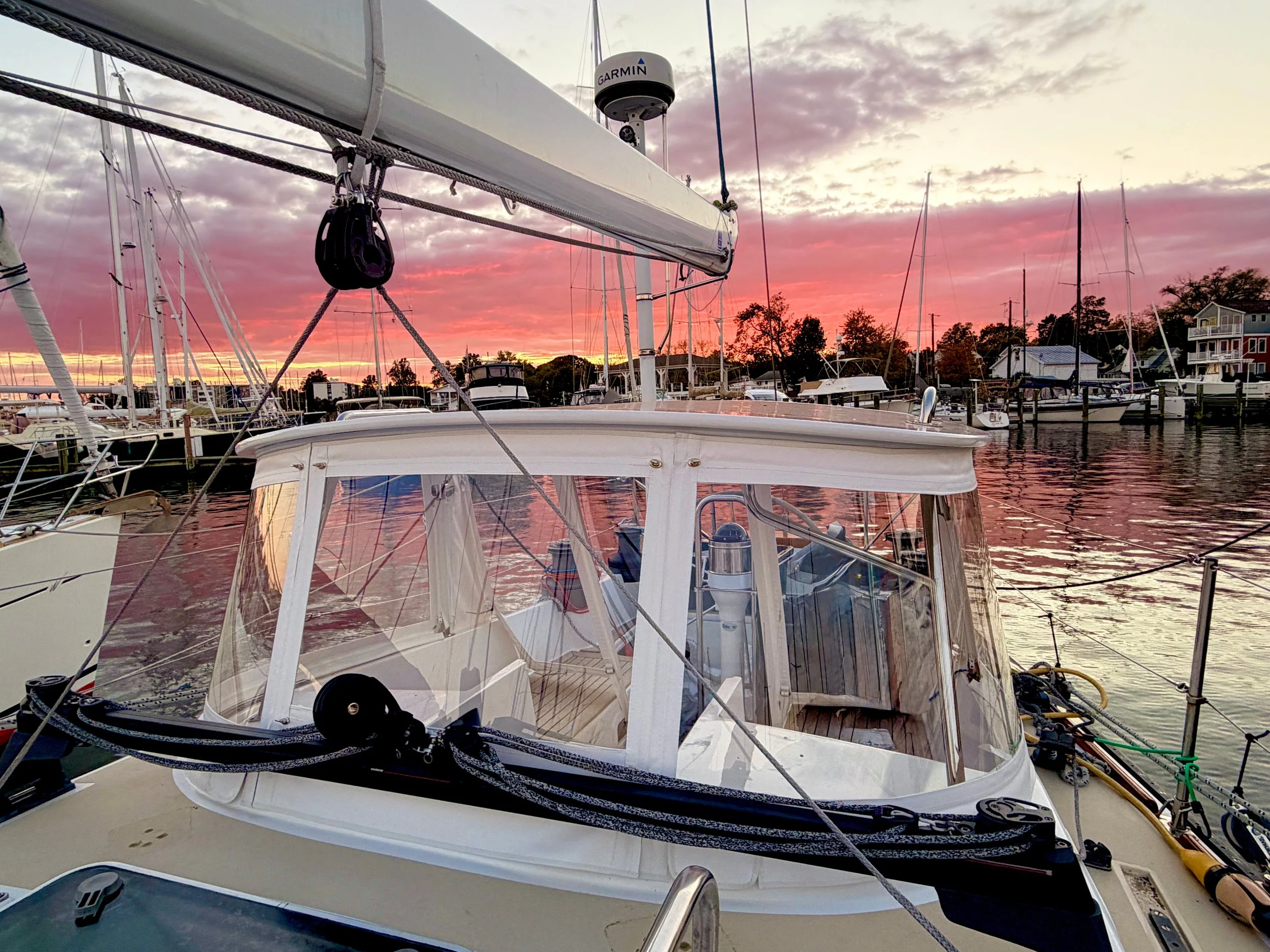 A sailboat docked at a marina during sunset with pink and purple clouds in the sky and other boats in the background.