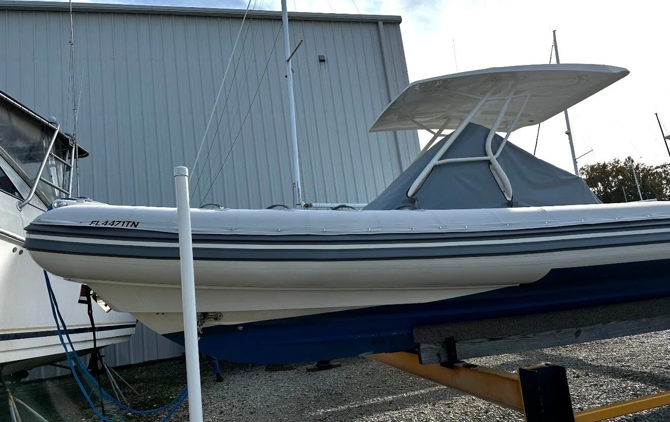 A white speedboat with a gray cover and a T-top, parked on a trailer in front of a gray industrial building.