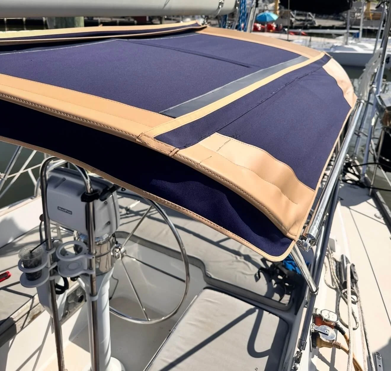Close-up of a boat's helm area showing a blue canopy with beige trim and a steering wheel, with other boats and a marina in the background.